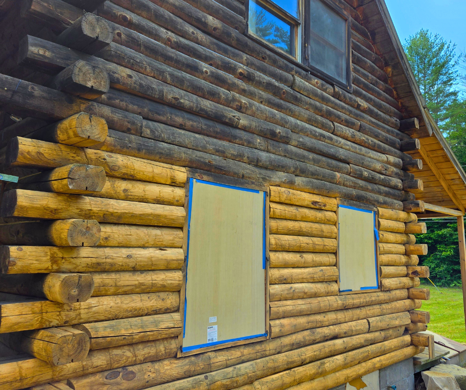 A log cabin under construction with two windows covered with cardboard and blue painter's tape on the beige lower part of the house. The dark upper logs of the cabin are visible, and the background shows green trees and a blue sky.