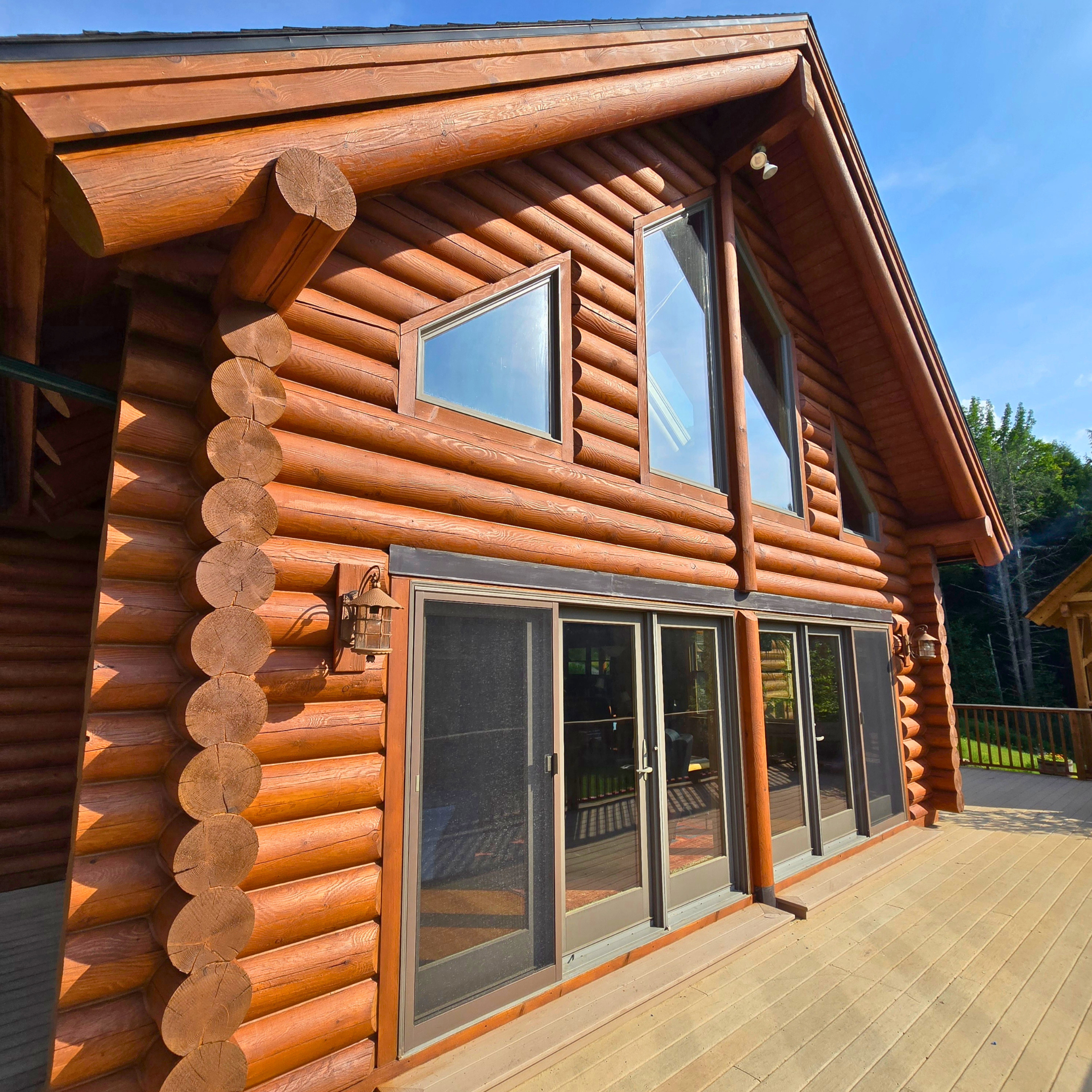 Exterior view of a wooden log cabin with large windows and sliding glass doors, situated on a wooden deck under a clear blue sky.