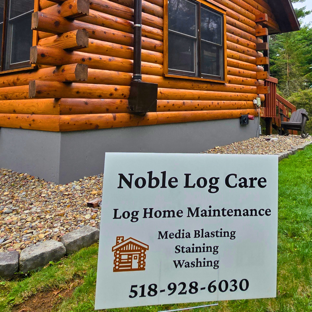 Sign for Noble Log Care showing services like log home maintenance, media blasting, staining, and washing, in front of a log cabin house with a gravel pathway, grass, and trees.