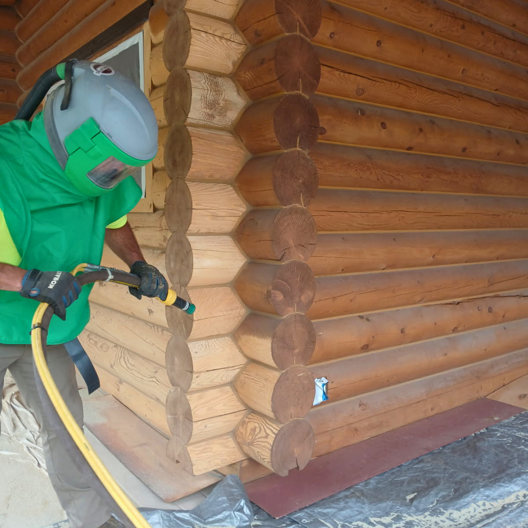 Person wearing a green shirt and protective gear using a pressure washer to clean the corner of a log cabin.
