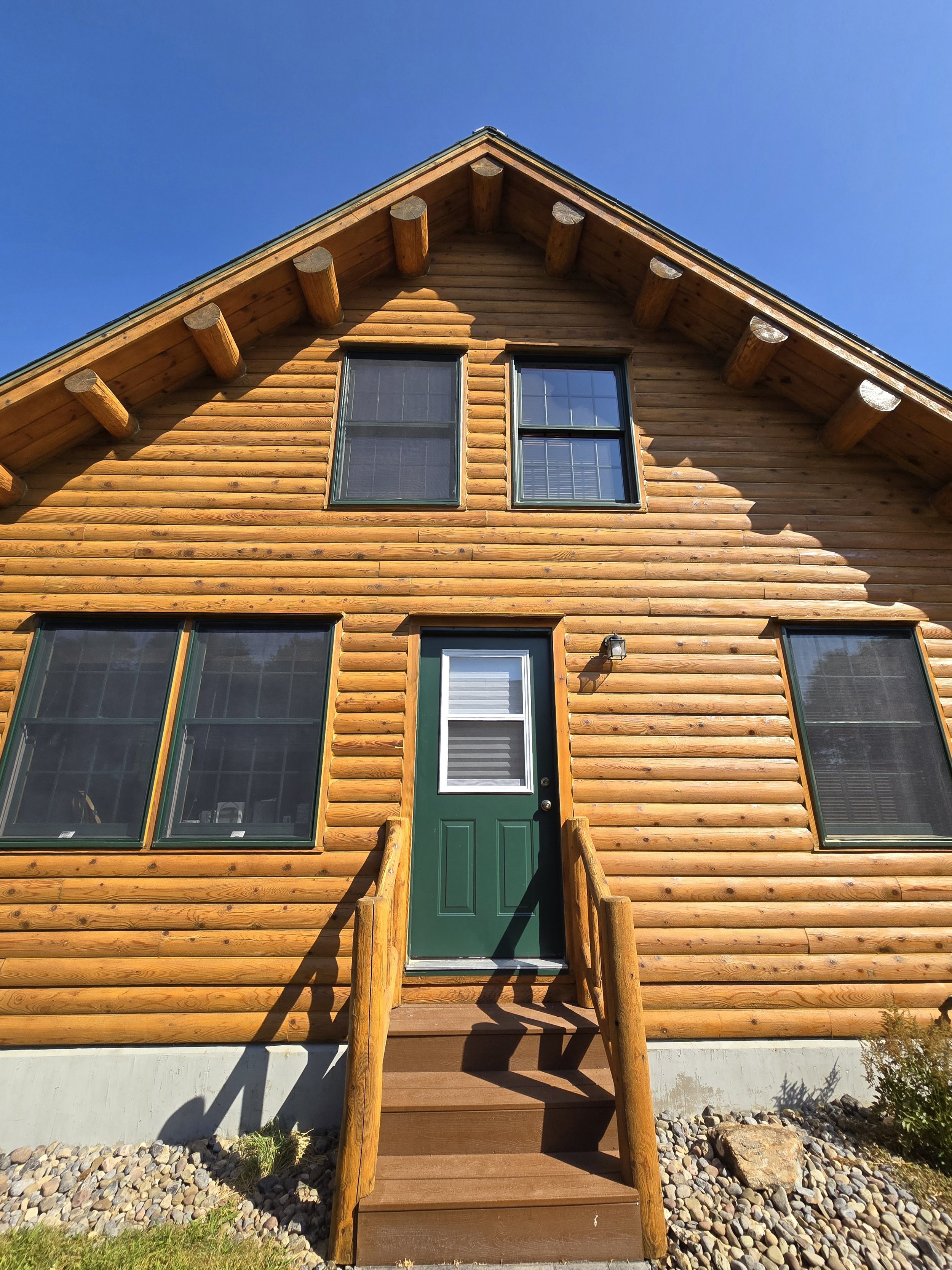 A wooden house with a steep roof, two central windows on the upper floor, four large ground-floor windows, a green door, and a small staircase with wood railings leading up to it. Rocks and plants are at the base of the house, with a blue sky overhea