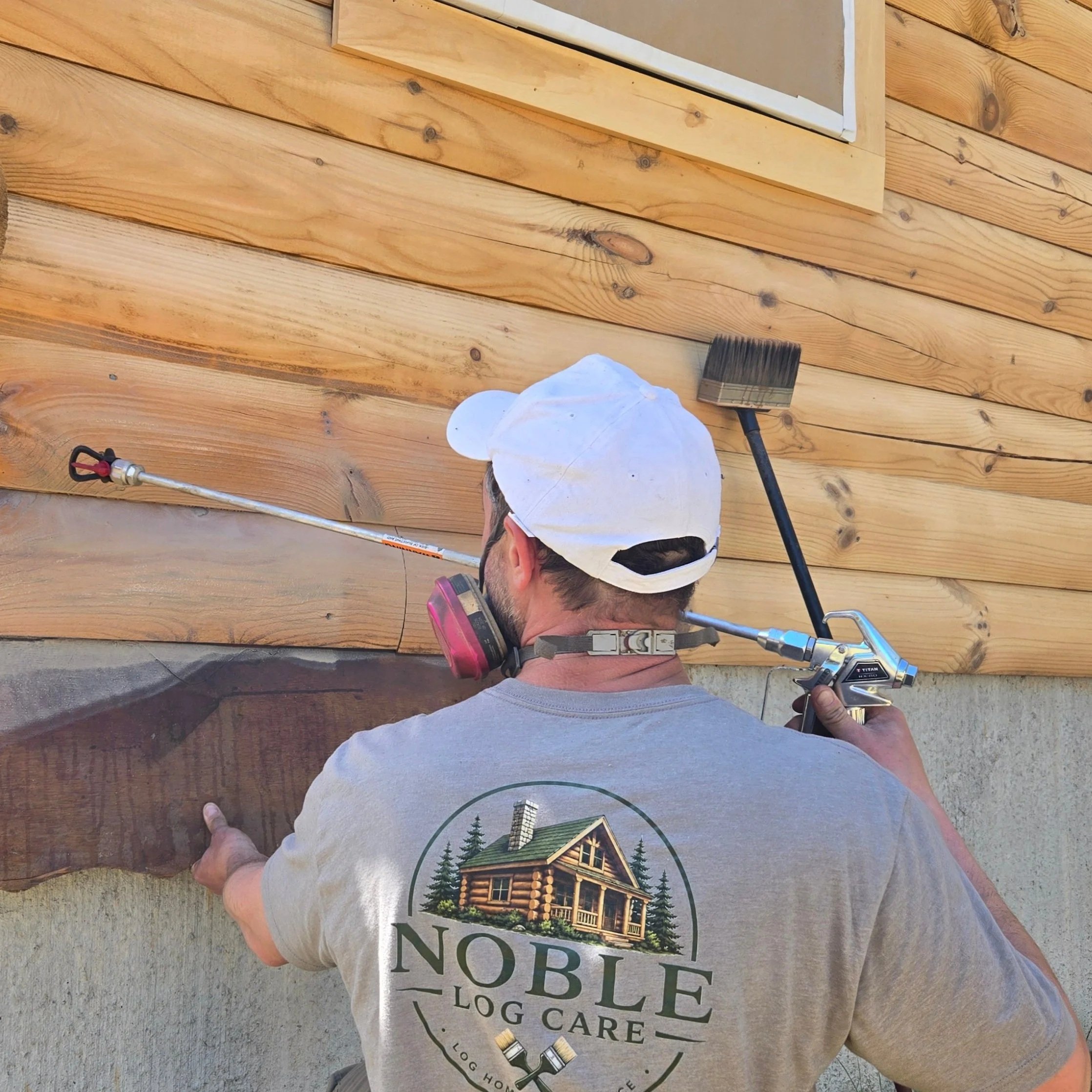 A man wearing a white baseball cap, facing away, holding a pneumatic nail gun on a wooden exterior wall, with a ladder in the background. The man is wearing a gray t-shirt with a logo of a log cabin and trees.