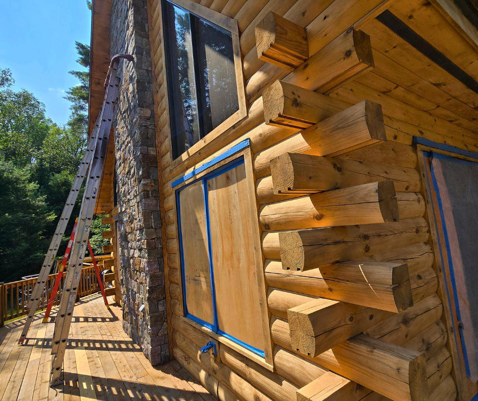 Close-up of a log cabin under construction with a stone chimney, a window, and a wooden deck; a ladder is leaning against the cabin.