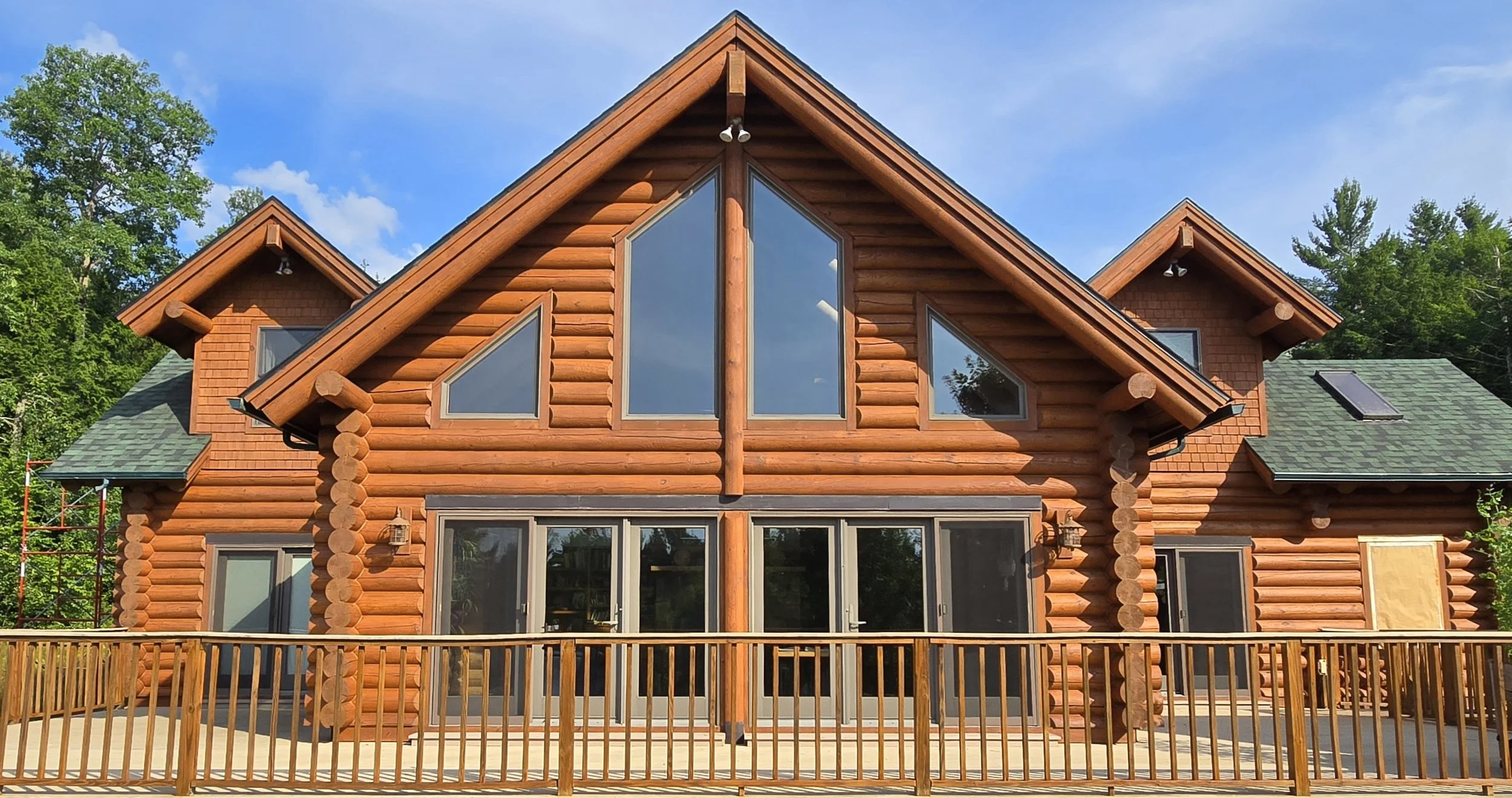 Front view of a large log cabin with pentagon-shaped windows in the upper center, a wooden railing in the foreground, and green trees and a blue sky in the background.