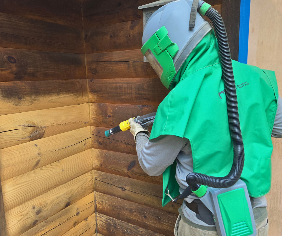 A worker wearing a helmet, green safety vest, and suit using a spray wand to treat or clean wooden wall panels.