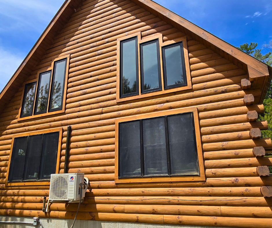 Front view of a wooden log cabin with several tall narrow and wide windows, an air conditioning unit, and a wooden roof, surrounded by greenery and a blue sky.