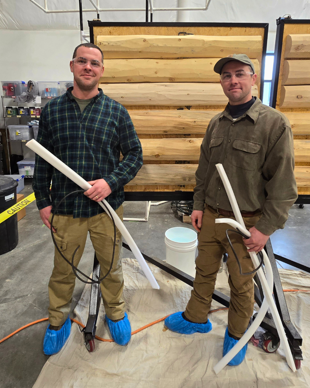 Two men in work clothes and safety glasses standing indoors on a tarp with construction supplies around them. They are holding large white tubes, wearing blue foot covers, with a background of wooden panels and a bucket.