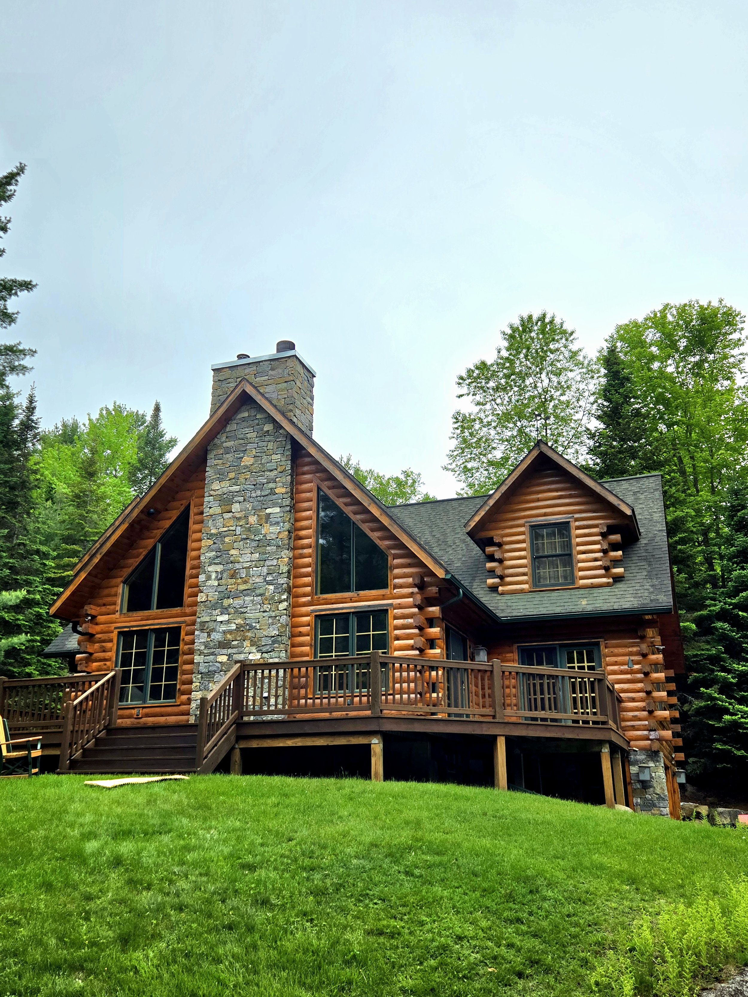 A rustic log cabin with a stone chimney, large windows, and a wooden deck surrounded by green trees and grass.