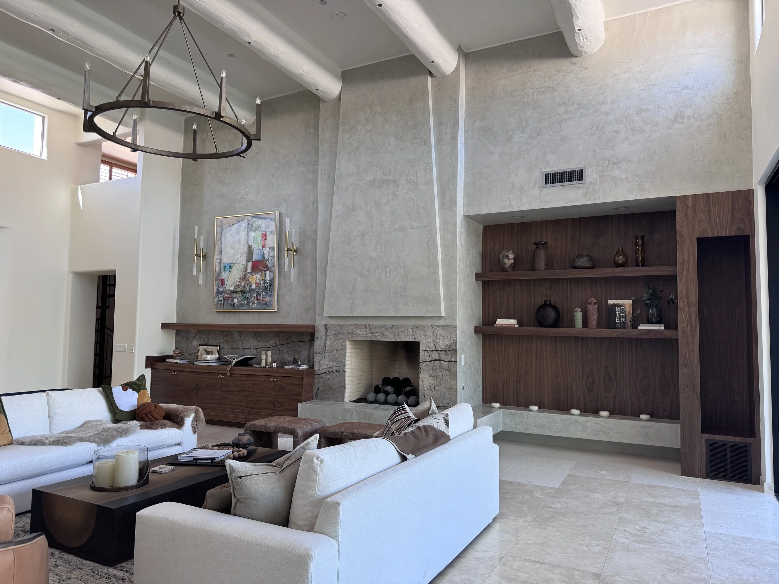 Modern living room with high ceiling decorated in neutral tones. Features a white sofa, coffee table with candles, artwork on the wall, and a fireplace with decorative vases in the built-in wooden shelf. Large windows allow natural light to illuminate the space.