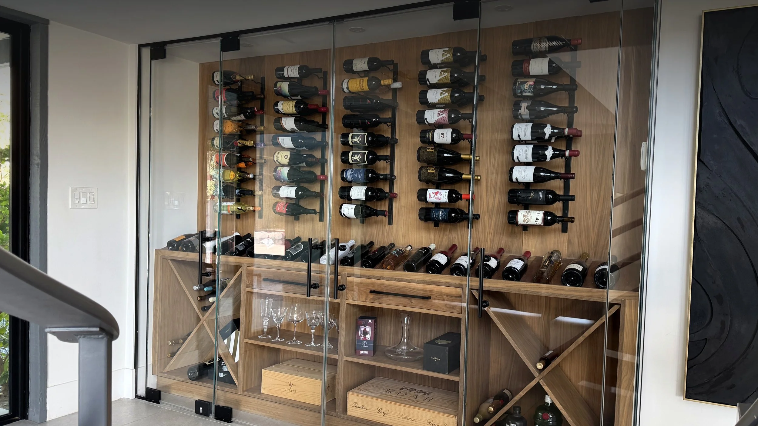 Glass-enclosed wine storage wall with horizontal shelving and a wood back panel beside a window.
