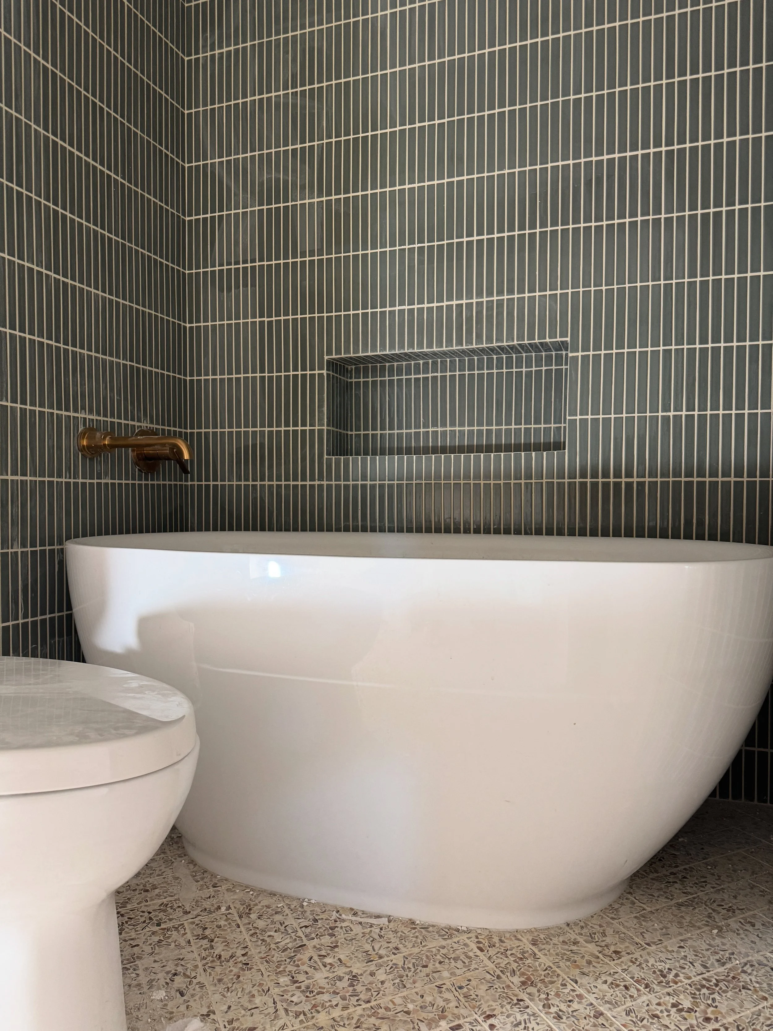 Renovated bathroom with a freestanding white tub, dark gray tile, and a patterned tile floor.