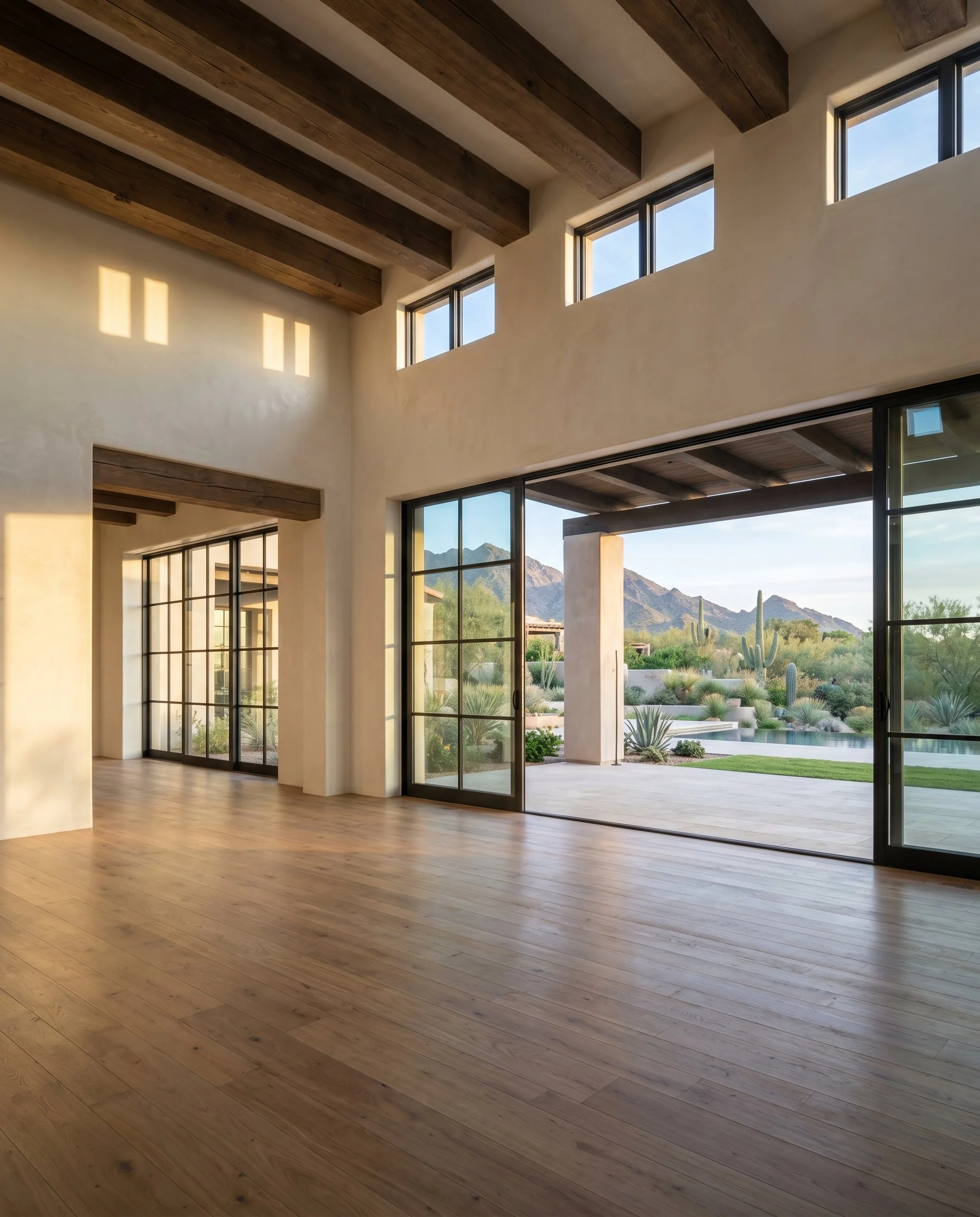 A modern interior space with wooden floors and ceiling beams, large glass windows, and a view of a desert landscape with mountains and cacti outside.