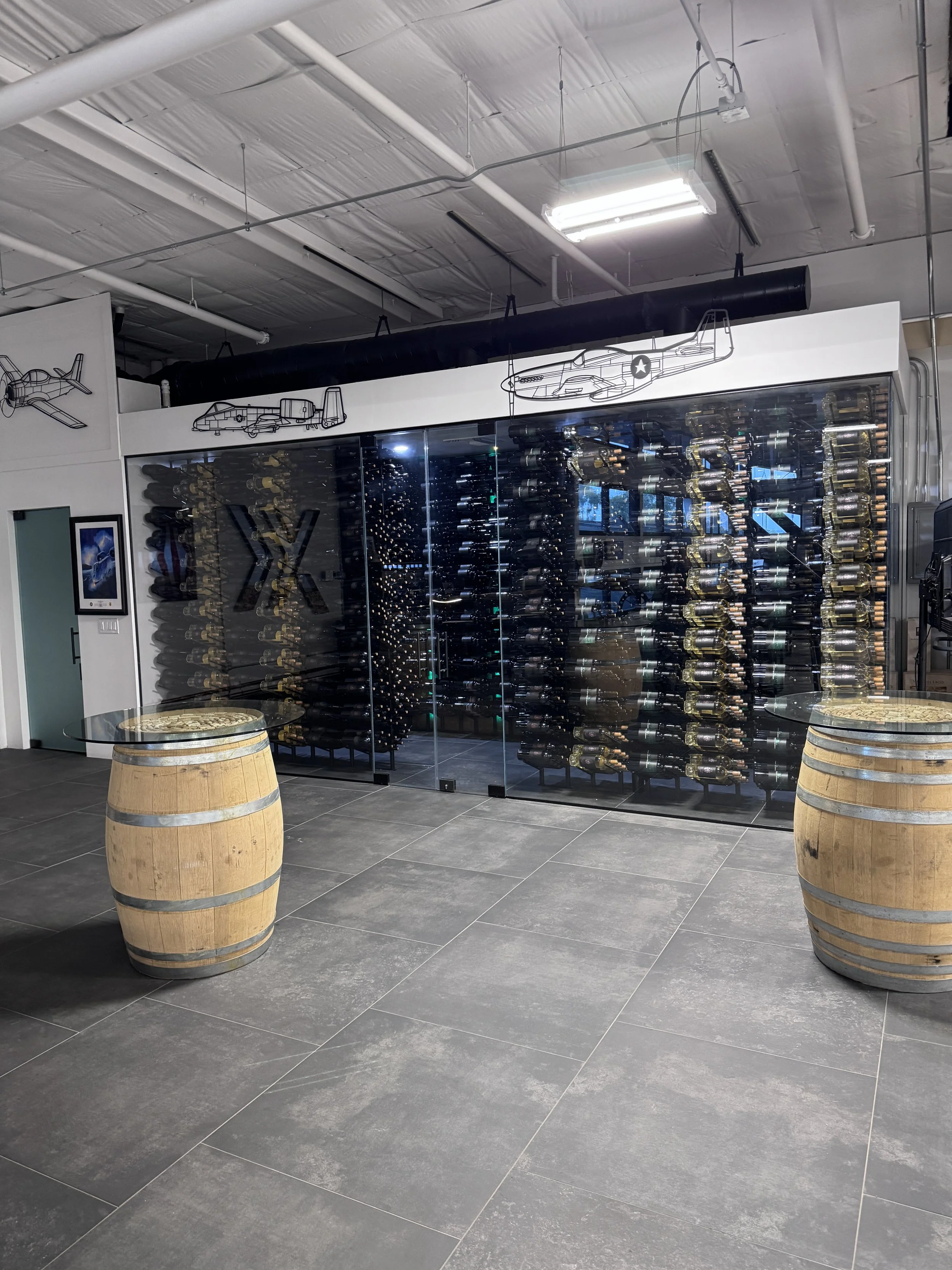 A wine store display with a glass wall filled with wine bottles, with aircraft artwork and a large black pipe on the ceiling, flanked by two wooden barrels used as tables.