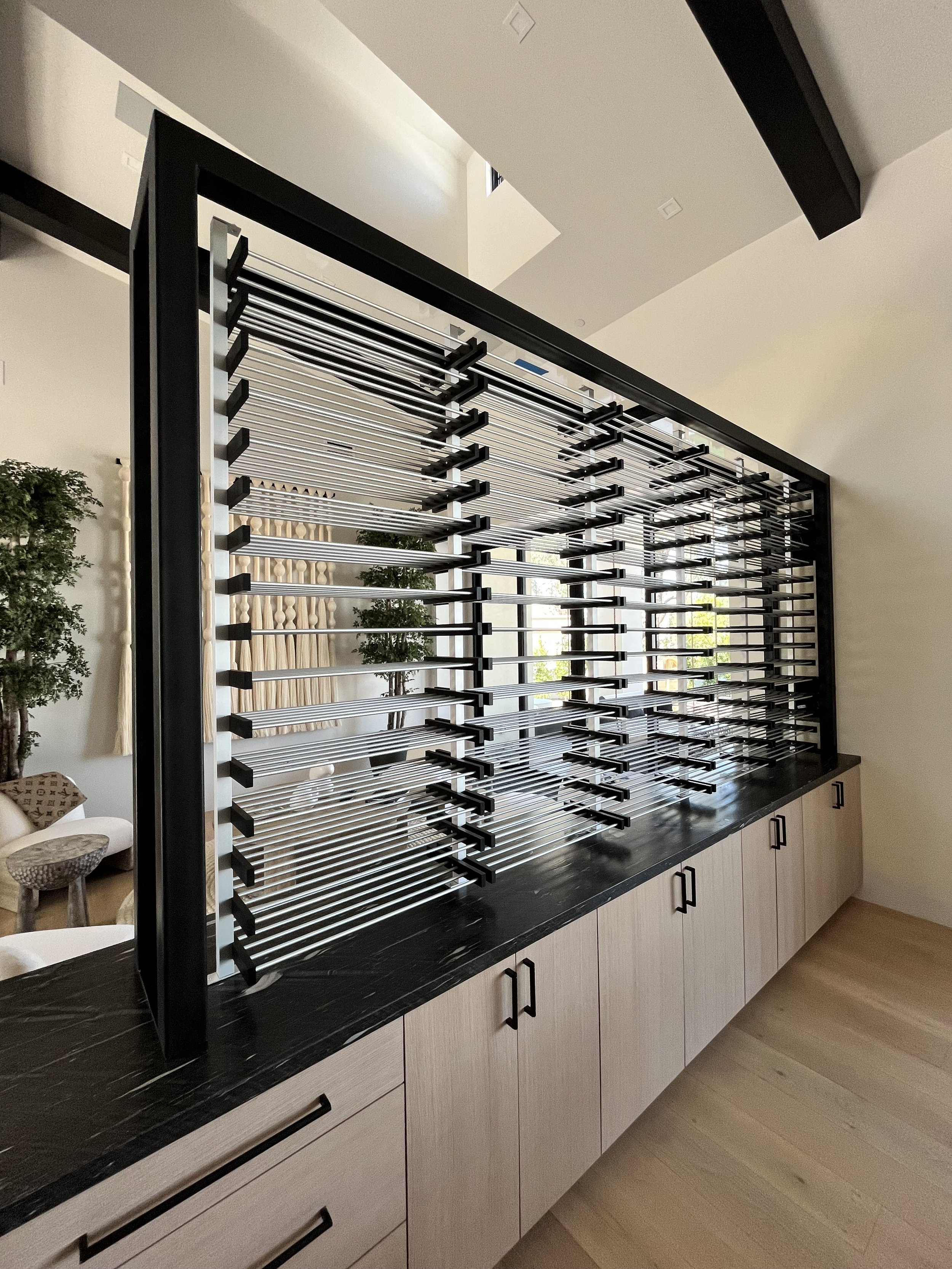 Wine display rack dividing two rooms, bottles stored horizontally on black metal pegs.