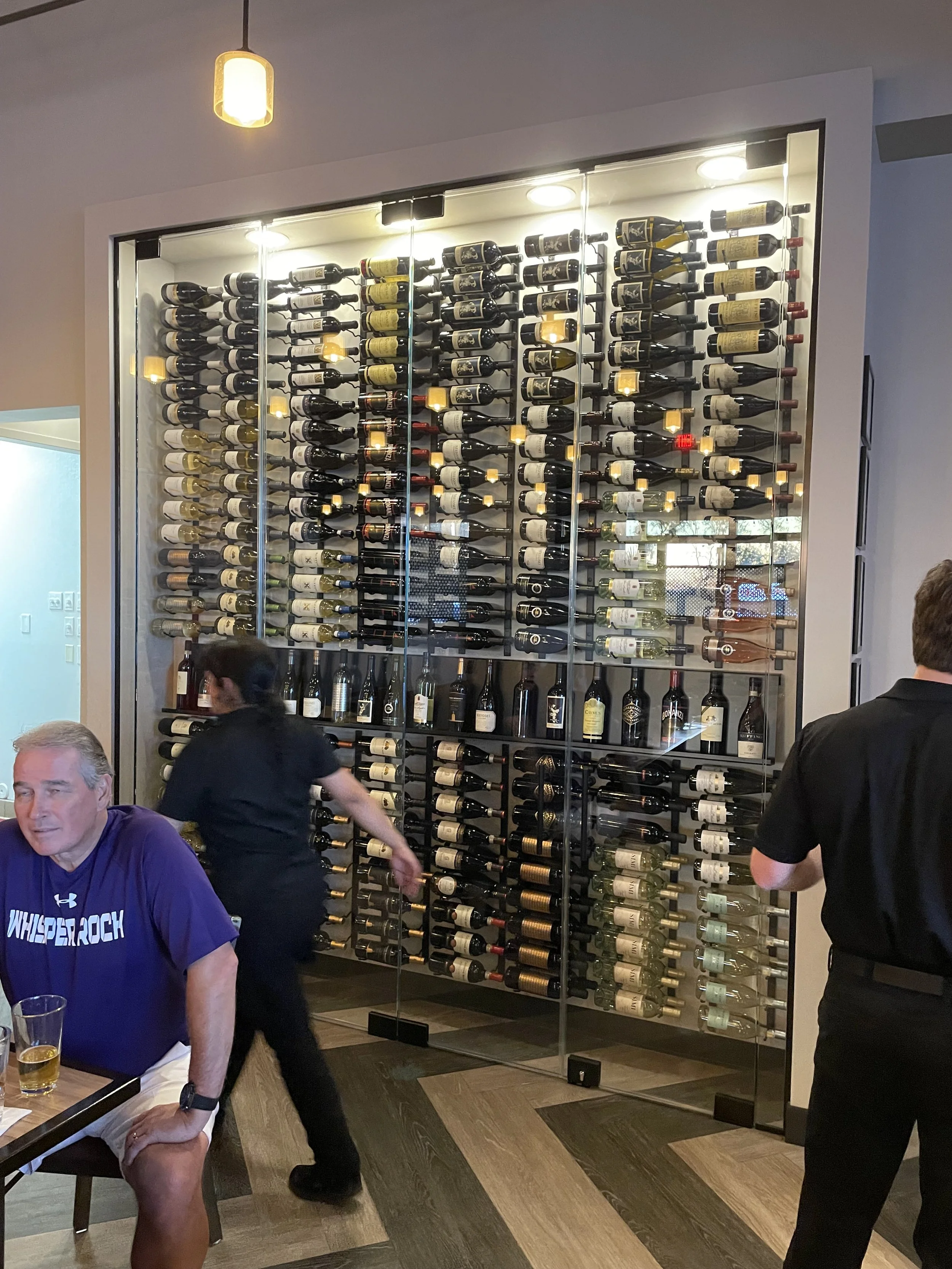 A wine rack with horizontal bottles behind glass doors in a restaurant or bar. Multiple people are present, with staff and a guest nearby