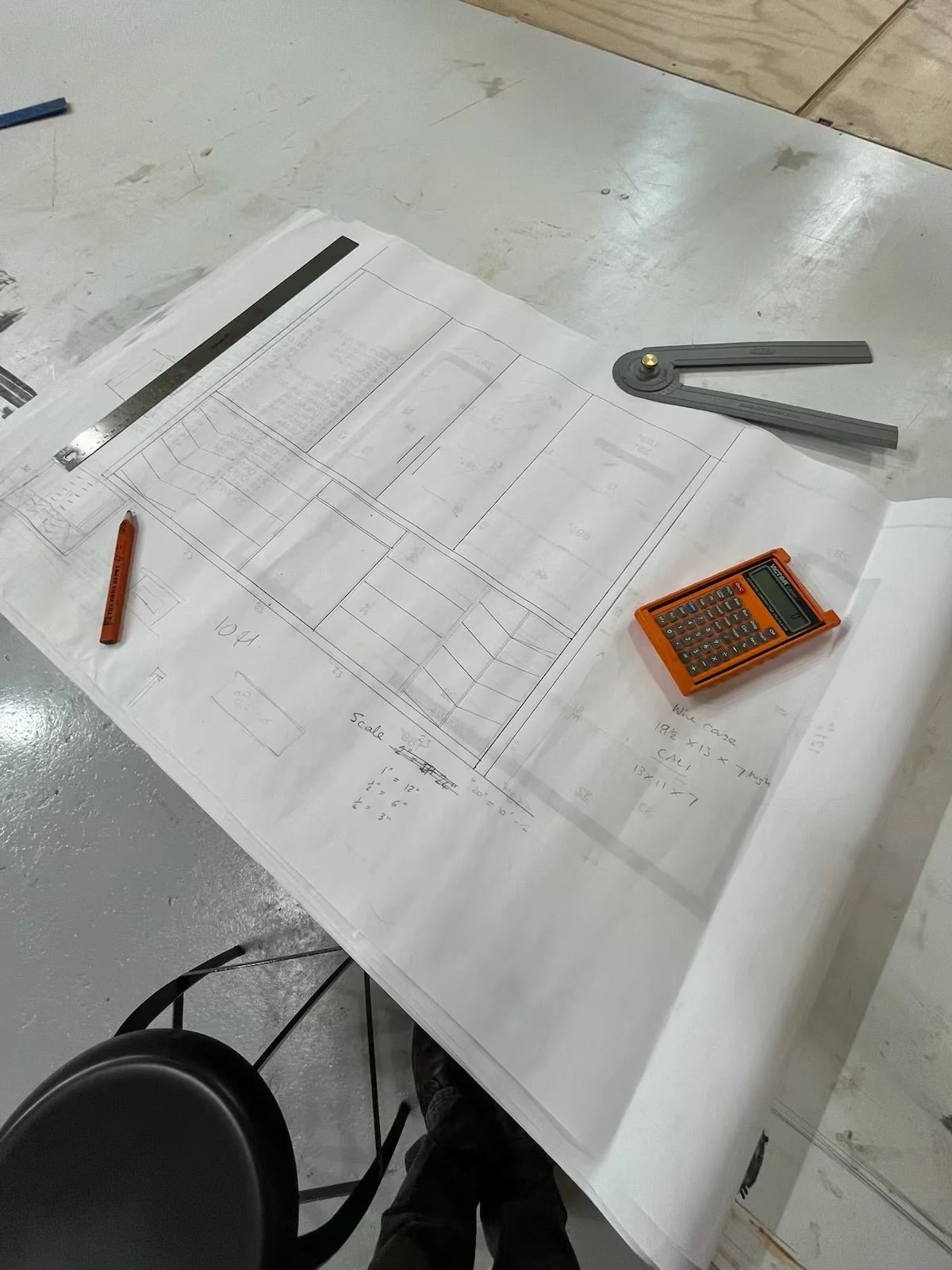 A construction or architectural workspace with a large blueprint on a table. On the blueprint, there is a metal ruler, a red marker, a small orange calculator, and a gray L-shaped adjustable drafting tool. The table has a plain white surface with some visible marks and stains.