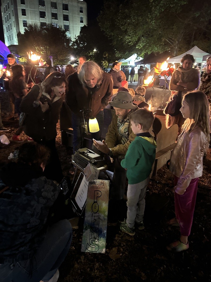 People gathered around a isaiah a typewriter poet seated with his typewriter on a bench at night, with a sign that reads ' Custom Poetry,' during a nighttime outdoor event with tents, trees, and a building in the background.