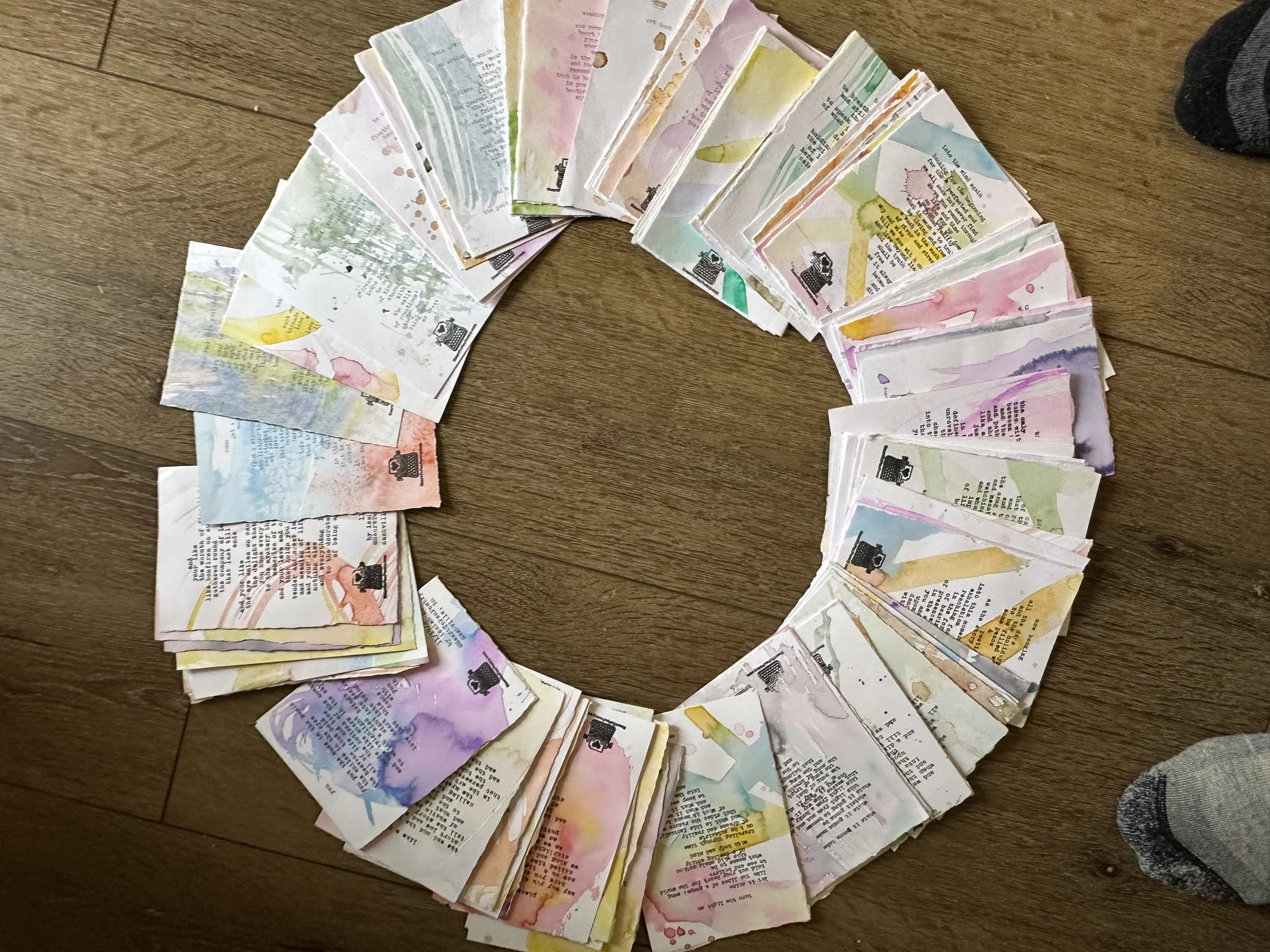 A circle shape outline made of several handwritten and watercolor-painted typewritten poems arranged on a wooden floor. The papers contain mostly text, with some colorful watercolor splashes.