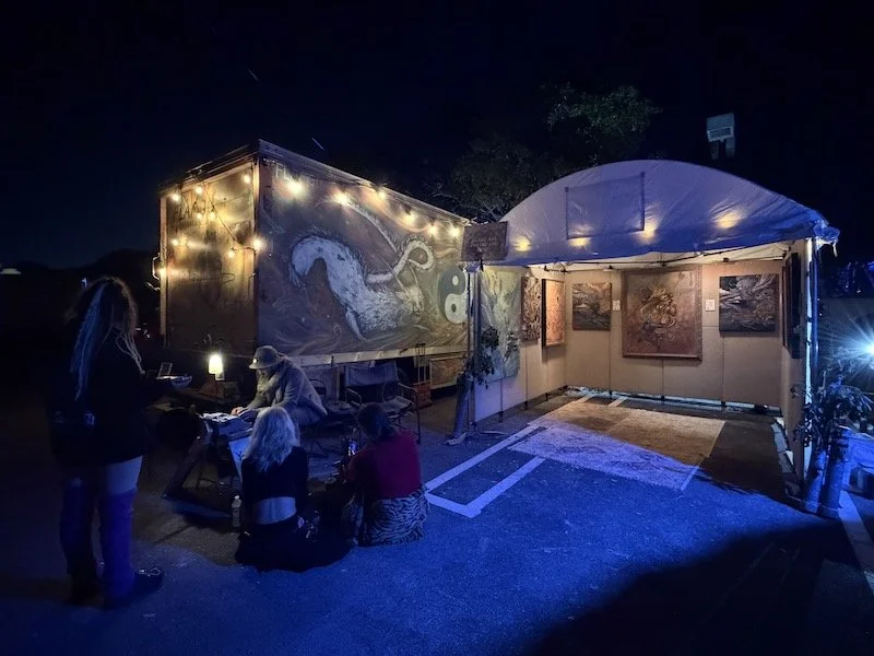 Outdoor art gallery at night featuring paintings and typewritten poems on display inside a tent and on a trailer decorated with string lights, with people viewing and sitting around listening to the typewriter poet read their poem.