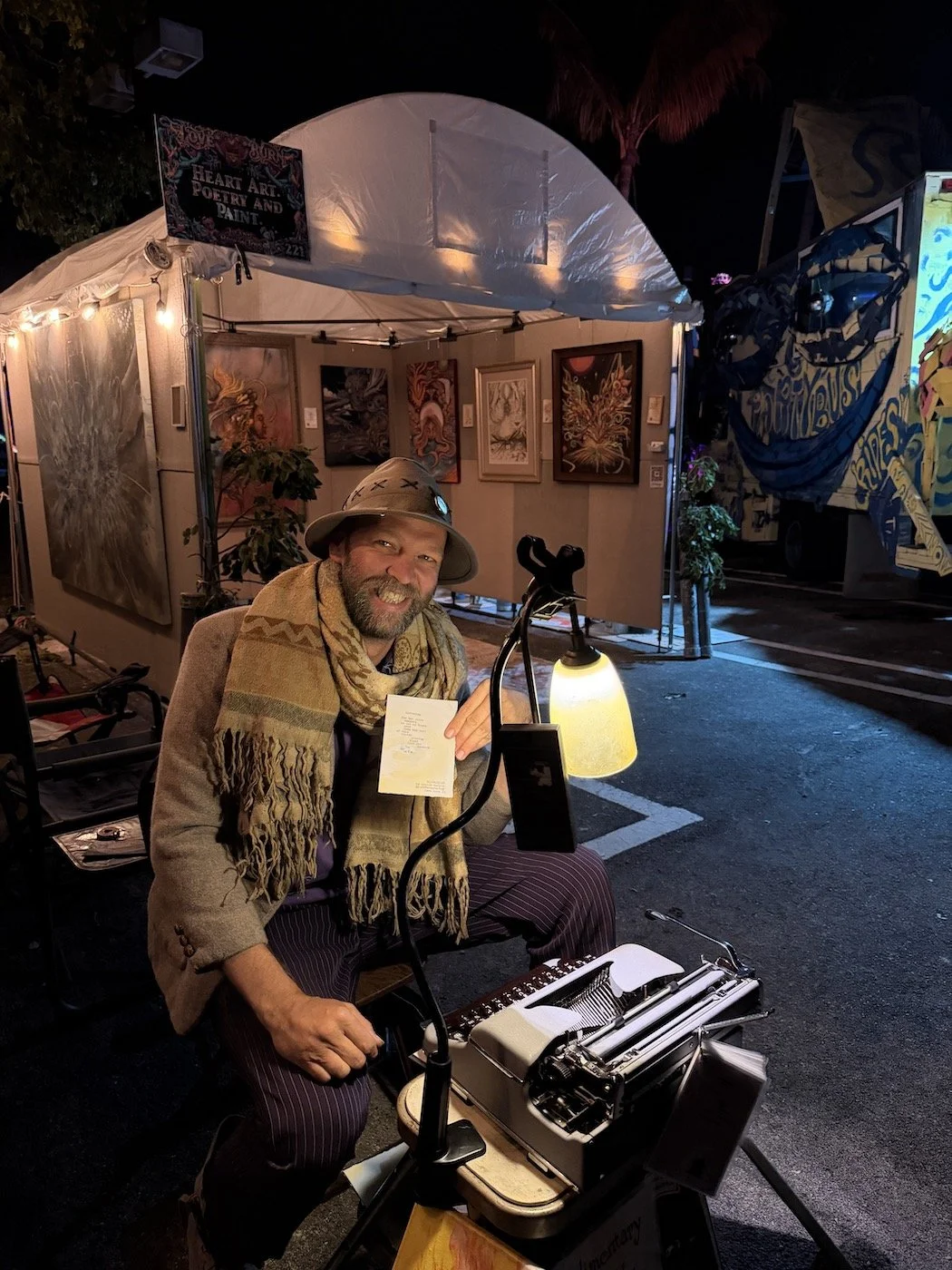 A man dressed in a vintage outfit, sitting outside a booth with artwork displayed on the walls. The man is smiling and holding a small piece of paper, with a vintage typewriter in front of him. The booth is part of an art event, with a sign that reads 'Heart Art Poetry and Paint,' illuminated by string lights at night.