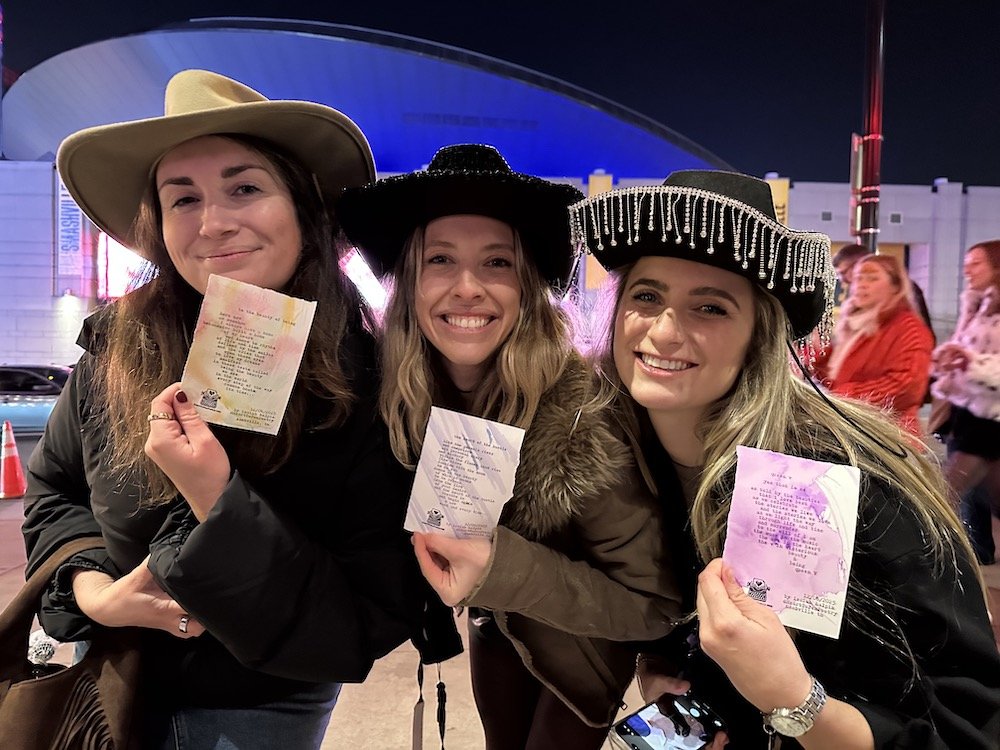 Three women wearing cowboy hats smiling and holding their typewriter poetry  on unique watercolored paper, standing outdoors at night with a modern building and other people in the background.