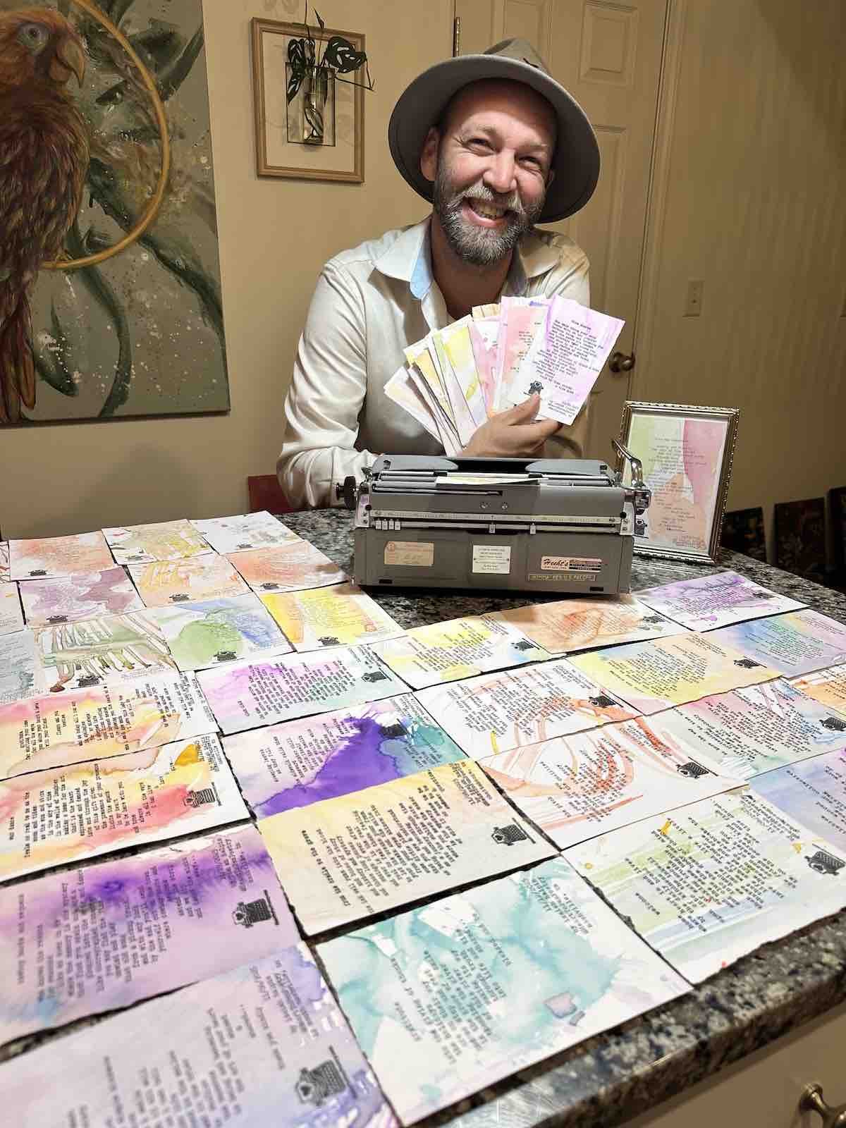 isaiah halpin is smiling and holding colorful typewritten and watercolor decorated poems. He is sitting with a typewriter  and a variety of personalized painted poems on the table each featuring  a one of a kind poem on watercolor.