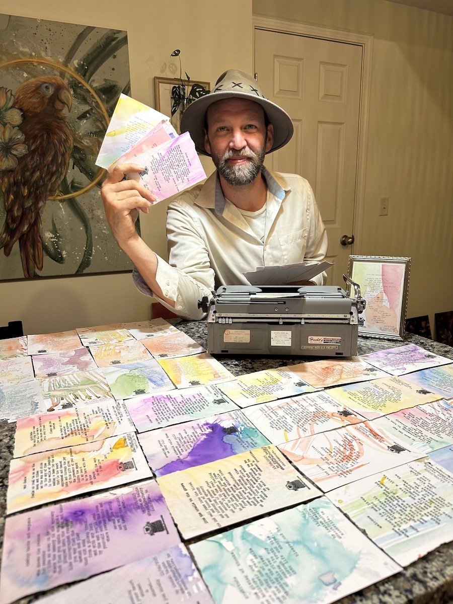 A man with a beard wearing a hat is sitting at a table with a vintage typewriter and colorful artwork or pages. He is holding some papers and smiling at the camera. Behind him is a painting of a bird and some framed pictures.