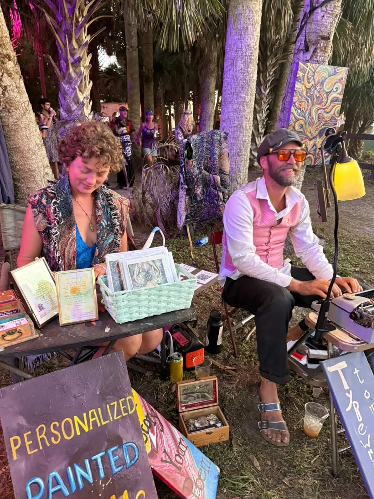 Two people sitting outdoors at a booth under palm trees, selling and displaying colorful poems artwork and paintings; Flamgu is live watercolor painting and isaiah live typewriting a poem for participants.
