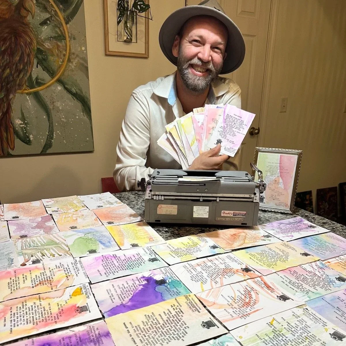 Isaiah Halpin, typewriter poet, with a beard, wearing a hat and a white shirt, smiling and holding colorful handwritten poems  in front of a vintage typewriter. The table is covered with similar colorful one of a kind typewritten poems