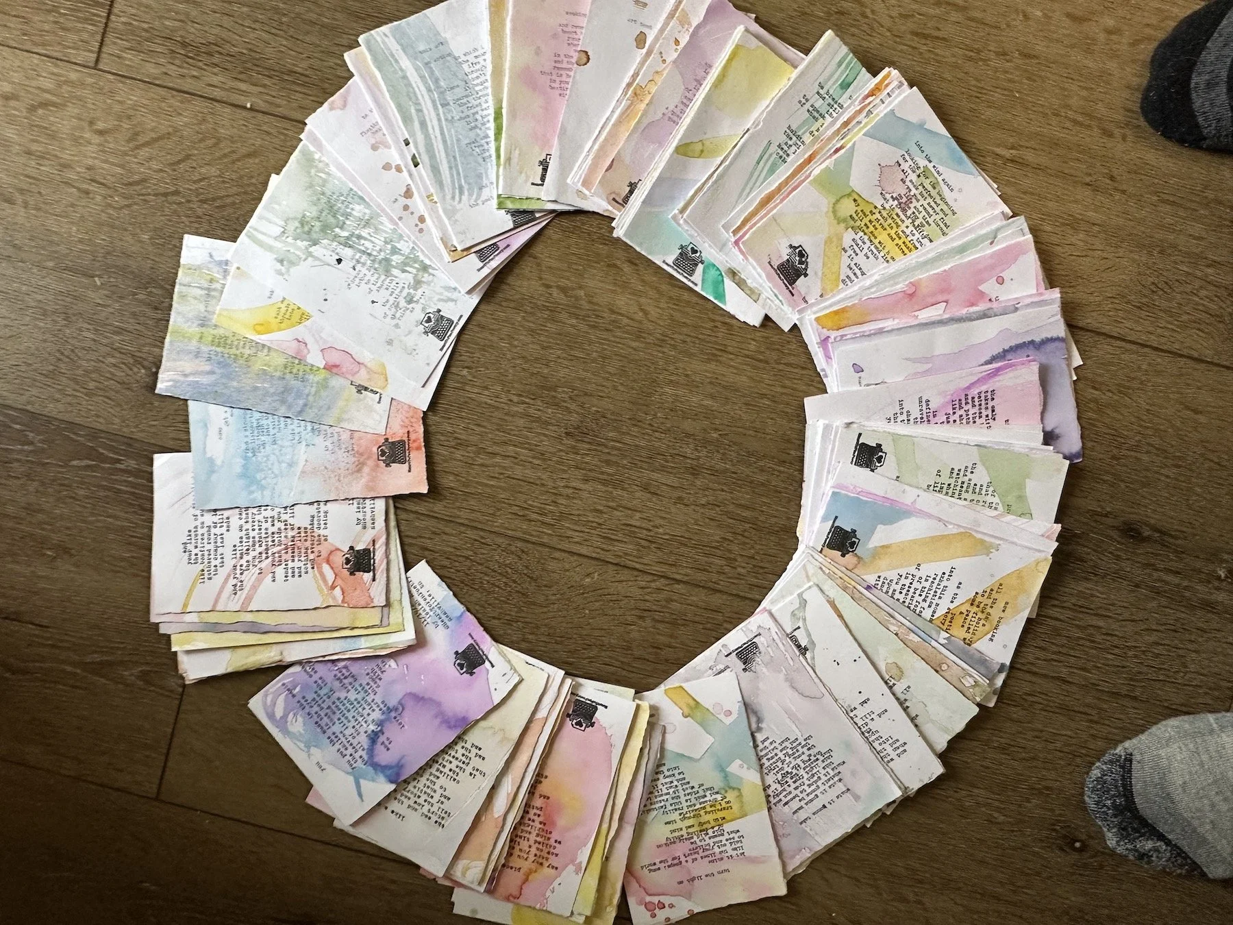 A circular arrangement of watercolor-painted typewritten poems, placed on a wooden floor.