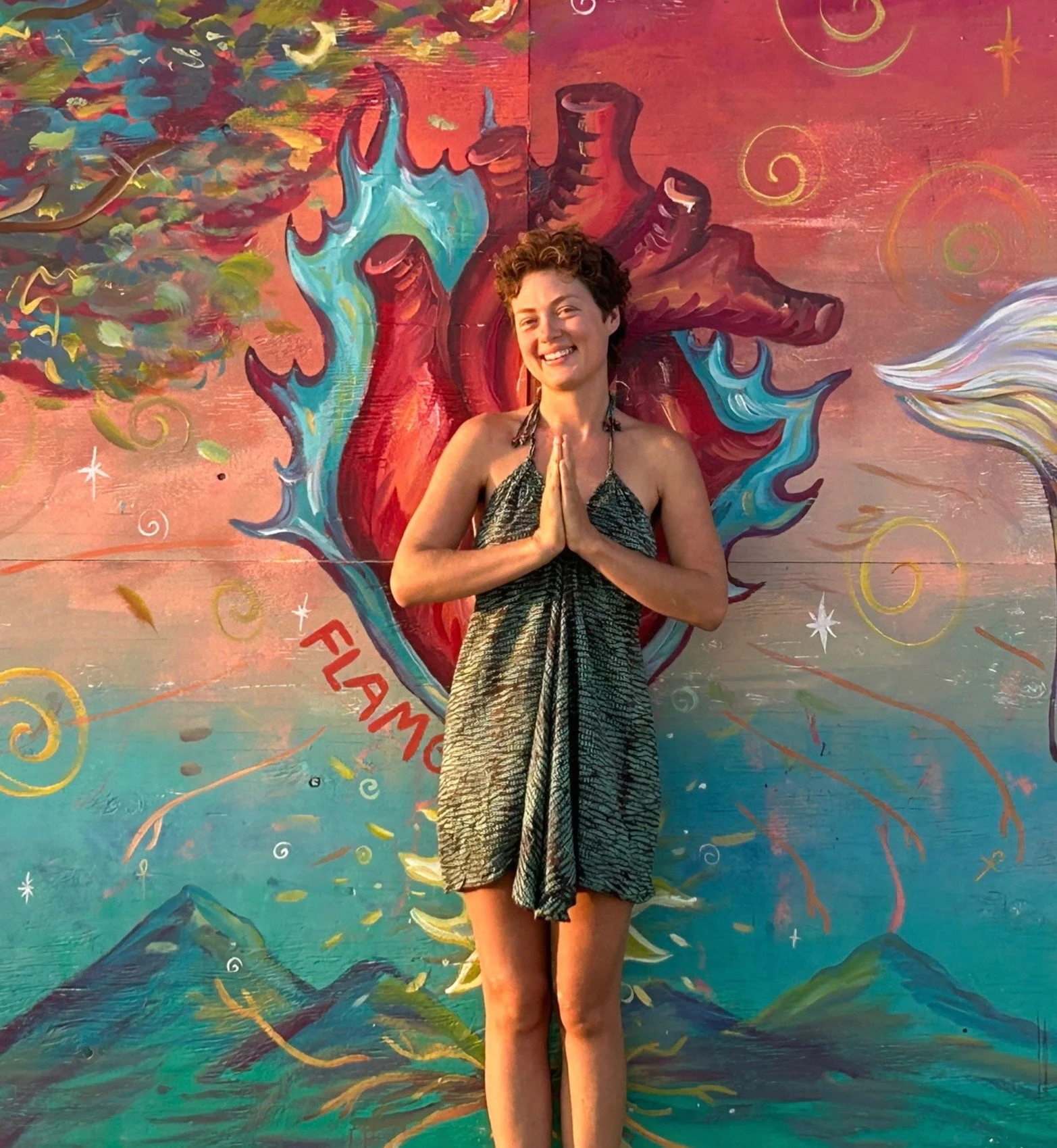 the artist Flamgu with short curly hair smiling and standing with her hands pressed together in front of a colorful mural of a heart surrounded by flames, stars, and swirls.