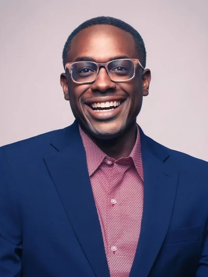 A smiling man wearing glasses, a blue suit jacket, and a pink patterned shirt against a plain light background.