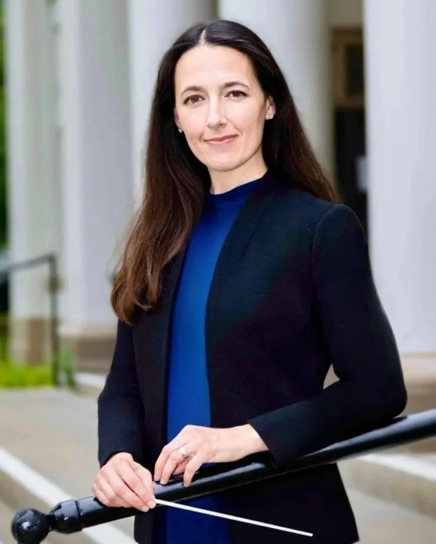 A woman with long brown hair, wearing a black blazer over a blue dress, holding a black baton, standing outdoors near a white building with greenery.