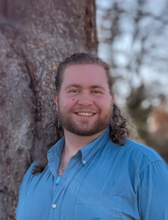 Man with long hair and beard smiling, leaning against a tree outdoors.