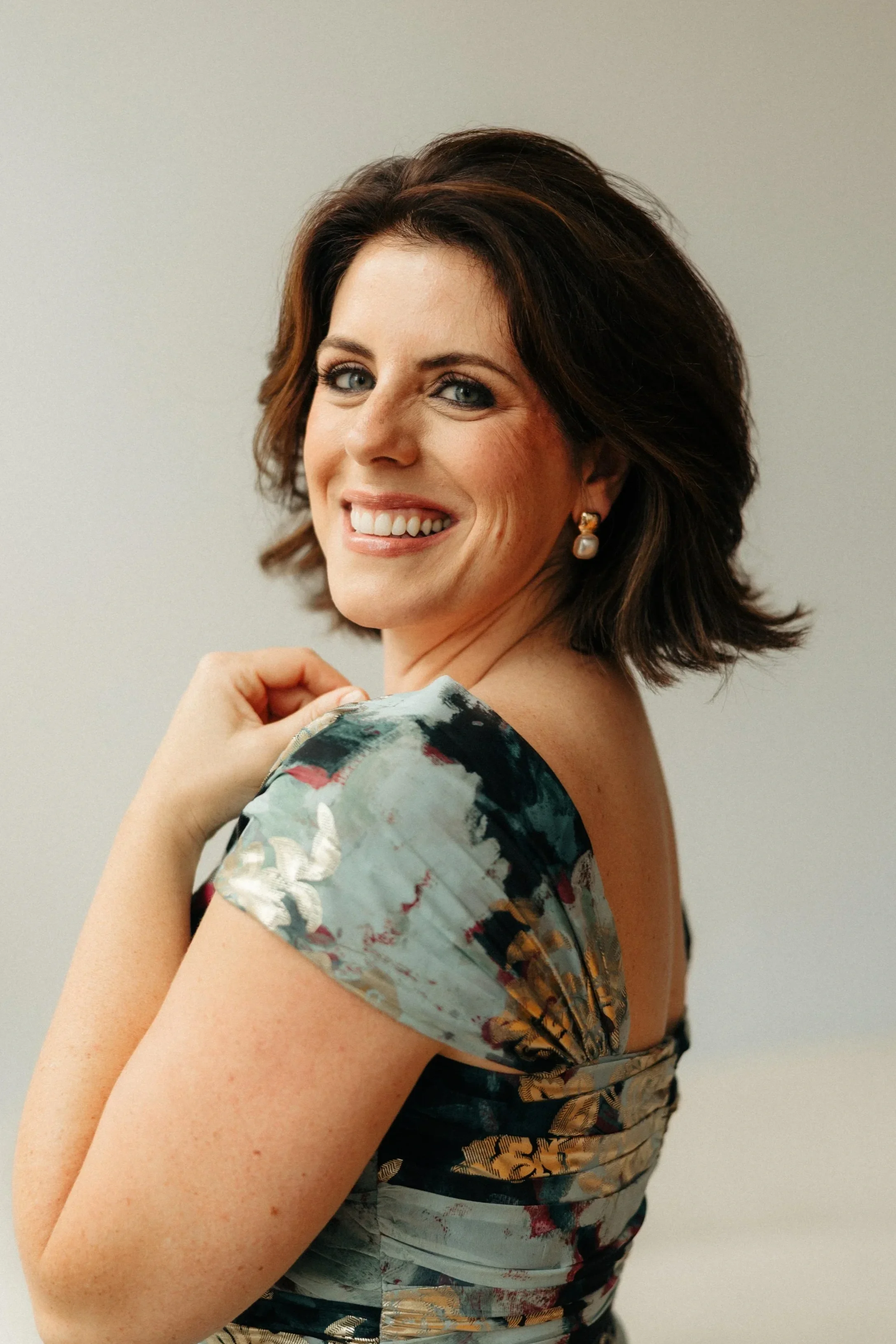 Close-up of a woman smiling, with short brunette hair, wearing pearl earrings and a floral dress, against a neutral background.