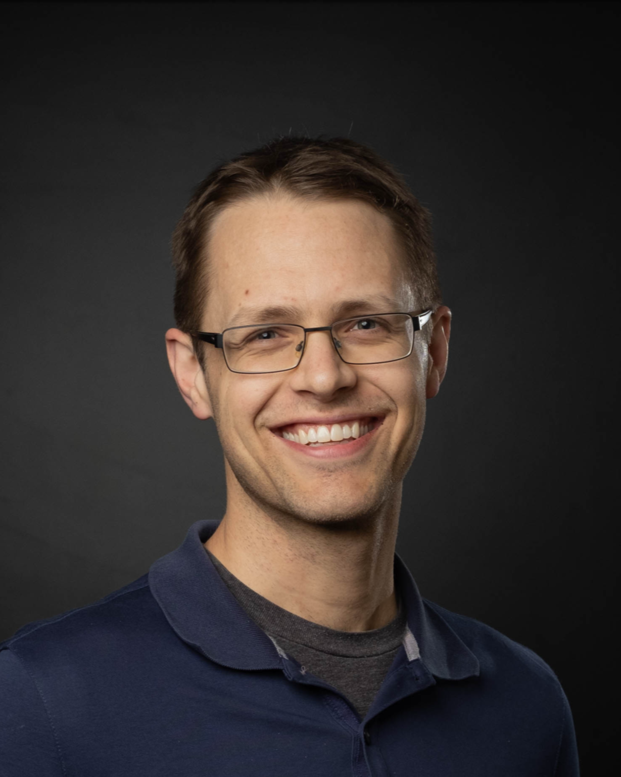 Portrait of a smiling man with glasses, wearing a dark blue collared shirt, against a dark background.