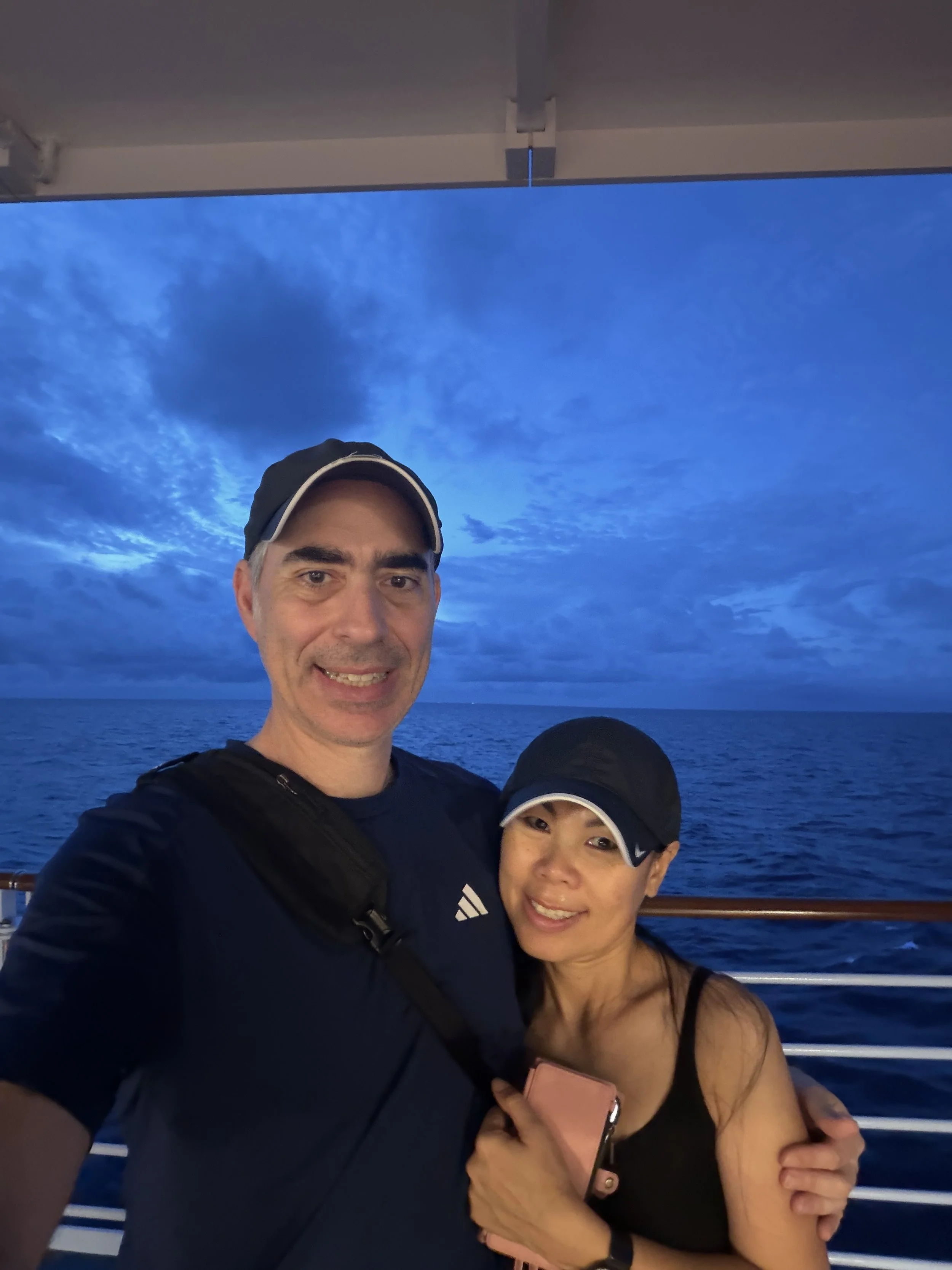A man and a woman taking a selfie on a boat during sunset. The sky is filled with blue clouds, and the sea is visible in the background.