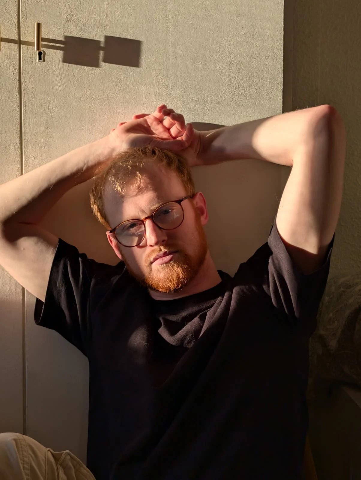 A man with glasses and a red beard is sitting on a couch with his arms behind his head, wearing a black shirt, against a beige wall.