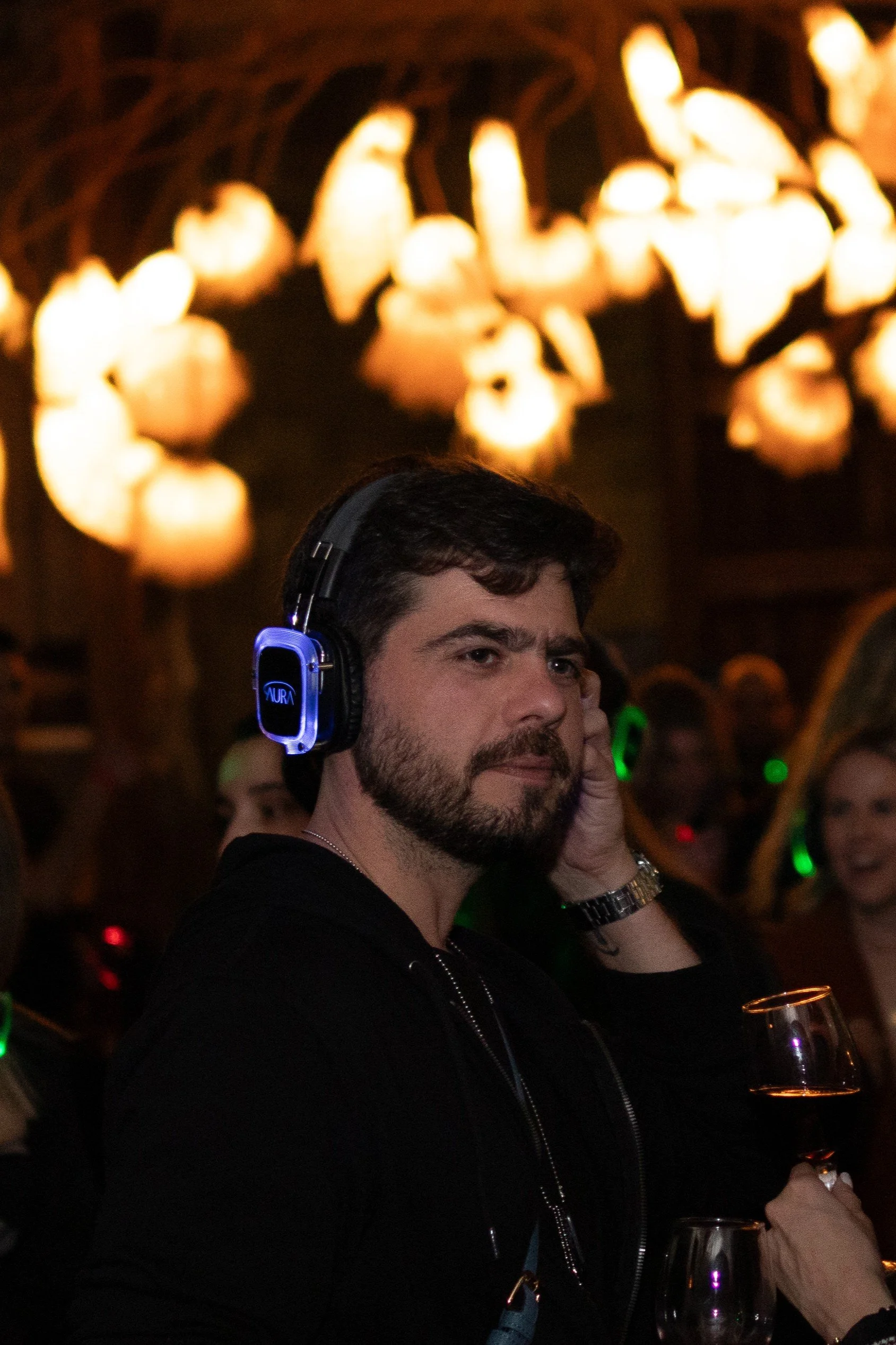 A man with dark hair and beard wearing headphones, holding a glass of wine, at a social gathering or party. Background features warm, glowing lights and other people.