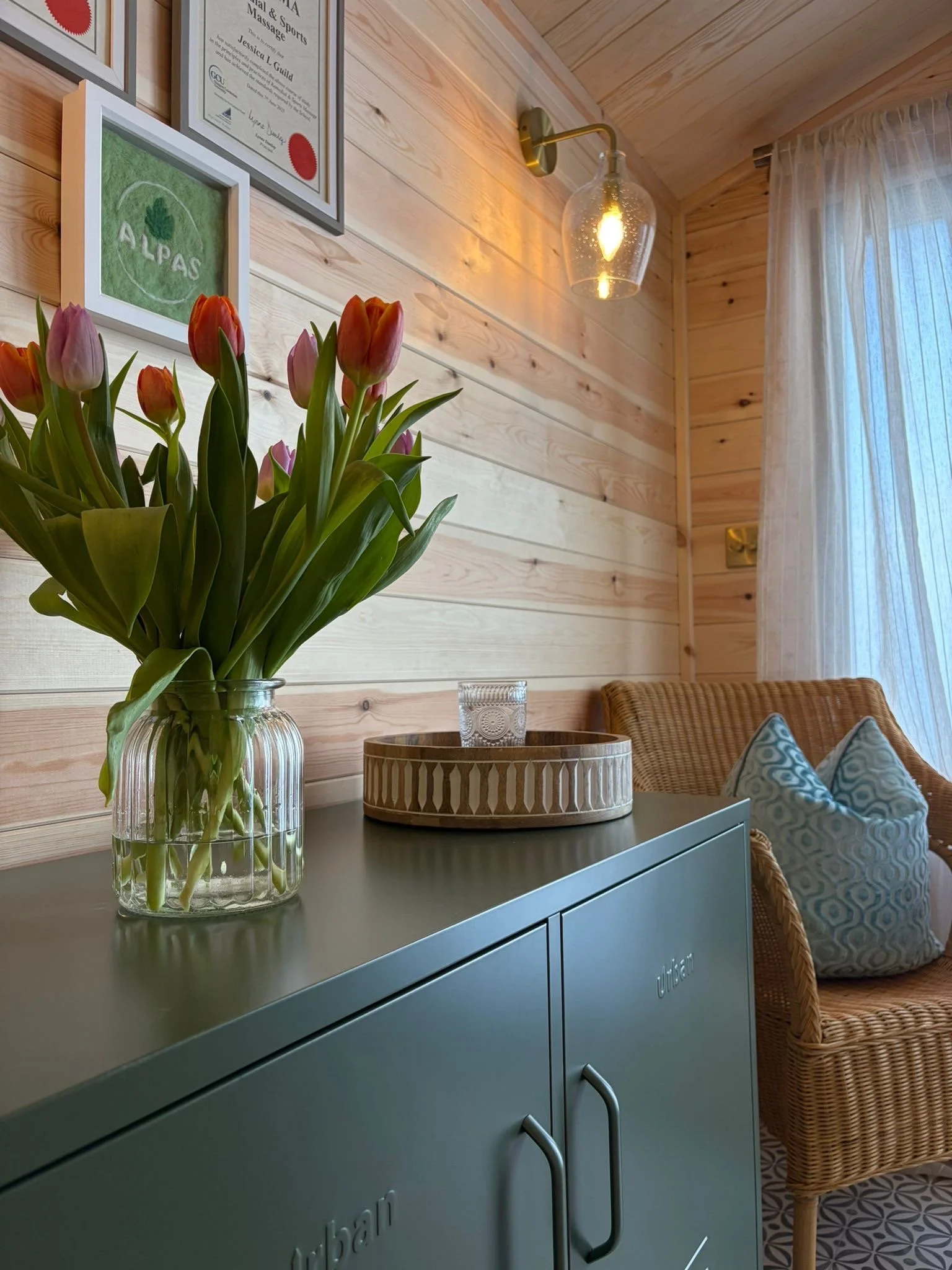 A colourful bouquet of pink tulips in a glass vase on a green cabinet. Behind, a textured wooden wall decorated with framed certificates and artwork, a wall-mounted light fixture, a window with sheer curtains, and a wicker chair with cushion.