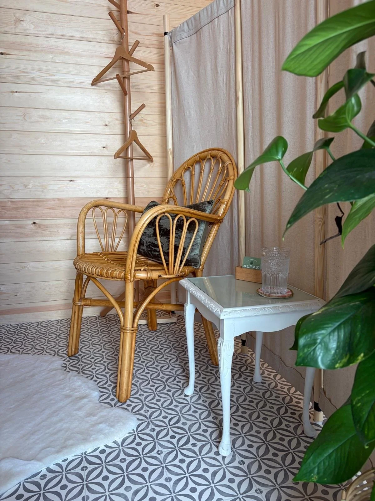 A cosy corner with a rattan armchair, a small white side table with a glass of water, a plant, and a wooden coat rack against a light wood-paneled wall and patterned tile floor.