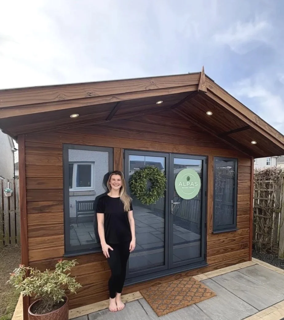 Business owner and massage therapist, Jessie, standing in front of the Alpas garden studio with glass double doors and a leafy wreath hanging on the door.