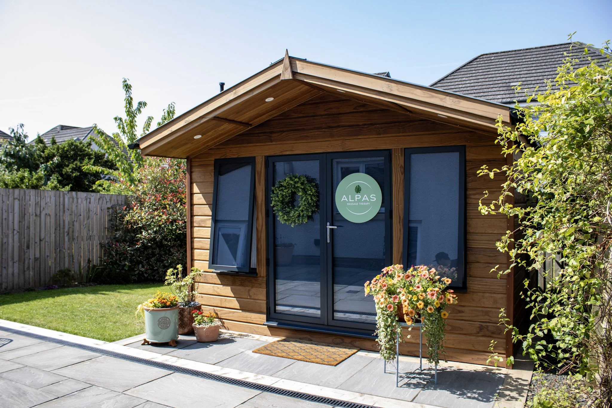 Wooden garden studio building with glass doors and a sign that says 'Alpas Massage Therapy'. There are potted plants and flowers outside, and a garden with grass and bushes surrounding the building. The sky is clear and blue.
