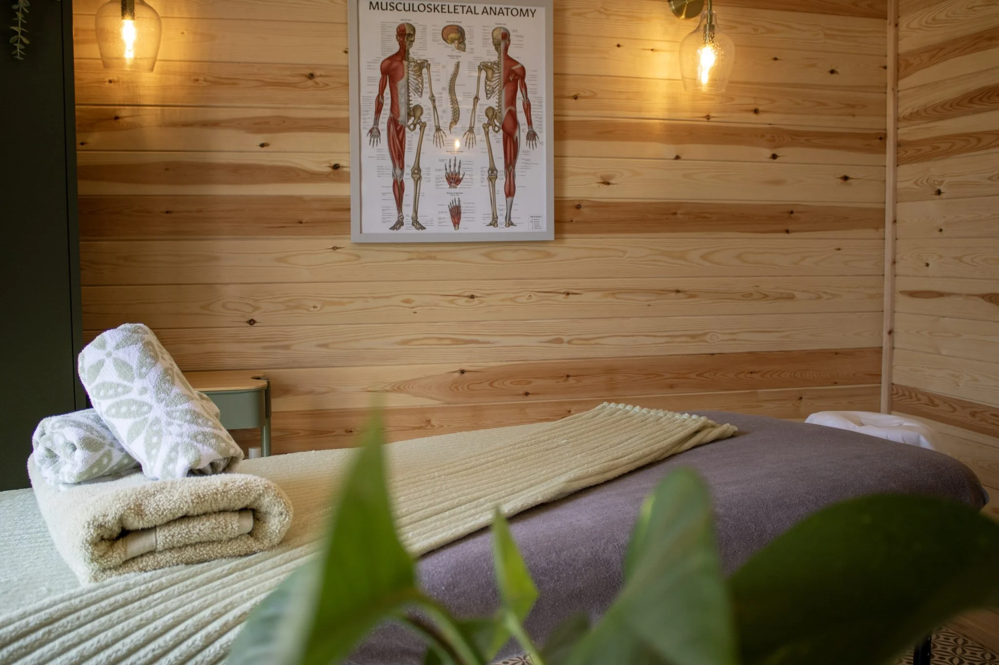 Treatment room with wooden panelled walls, two hanging pendant lights, a poster of muscular and skeletal anatomy, and a massage table with folded towels and a blanket.