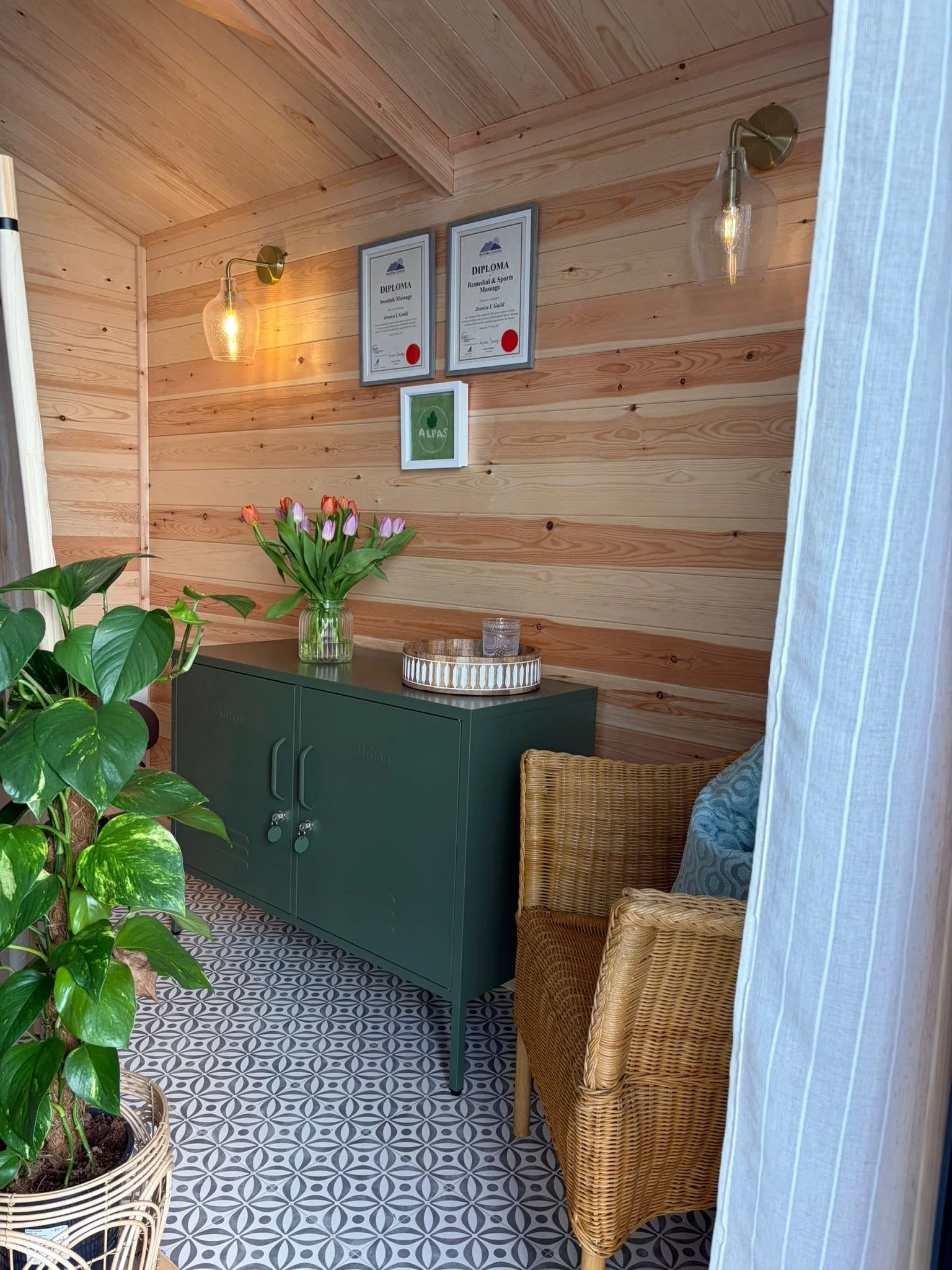 Interior of a cozy room with wooden walls and ceiling, featuring a green cabinet, framed diplomas, a table with a tray, potted plants, and woven chairs, illuminated by wall-mounted lights.