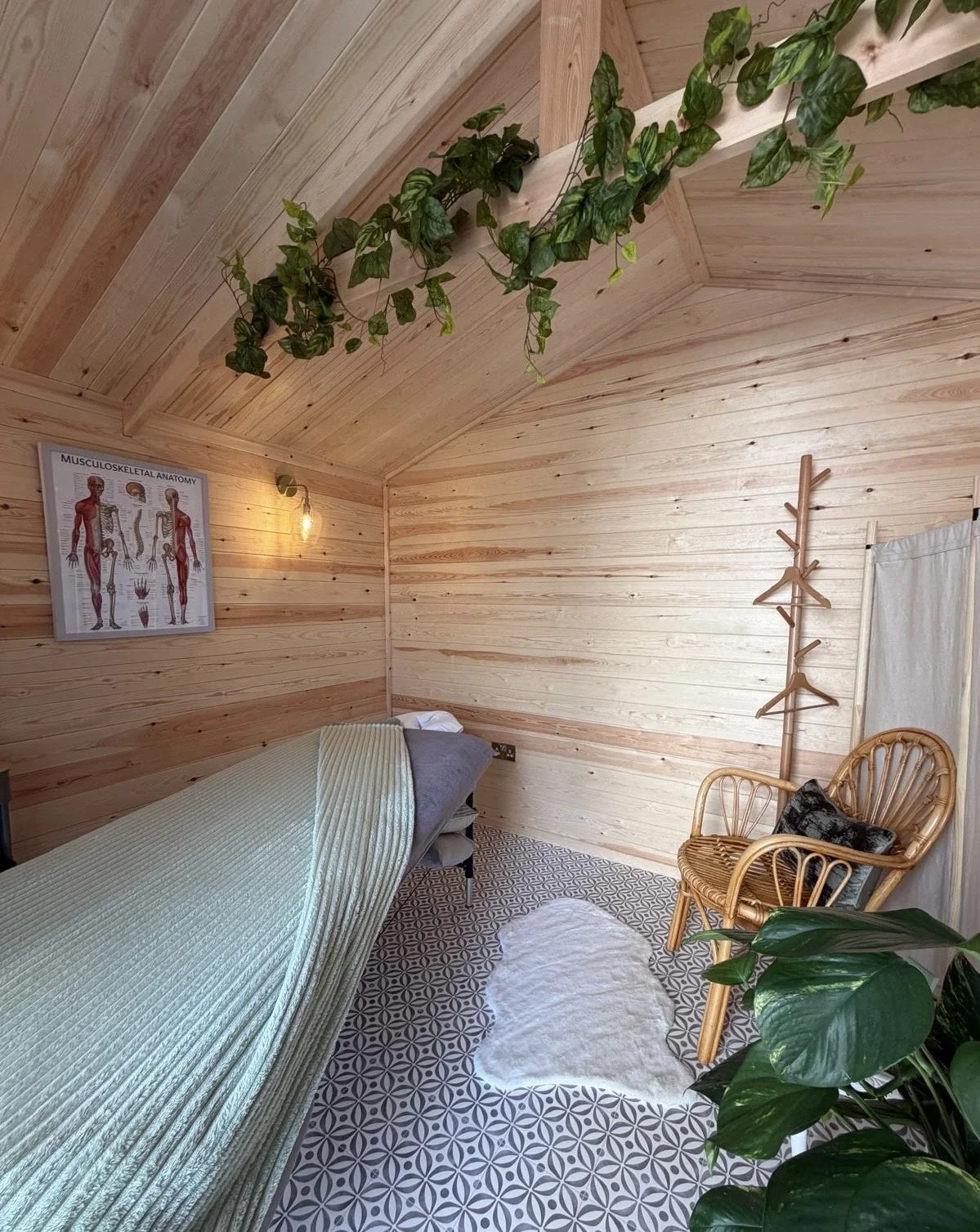 A cosy massage studio, featuring a massage table with blankets, a wooden coat rack, a rattan chair with cushion, a potted plant, a ceiling decoration of greenery, a wall-mounted light fixture, and a musculoskeletal anatomy poster on the wall.