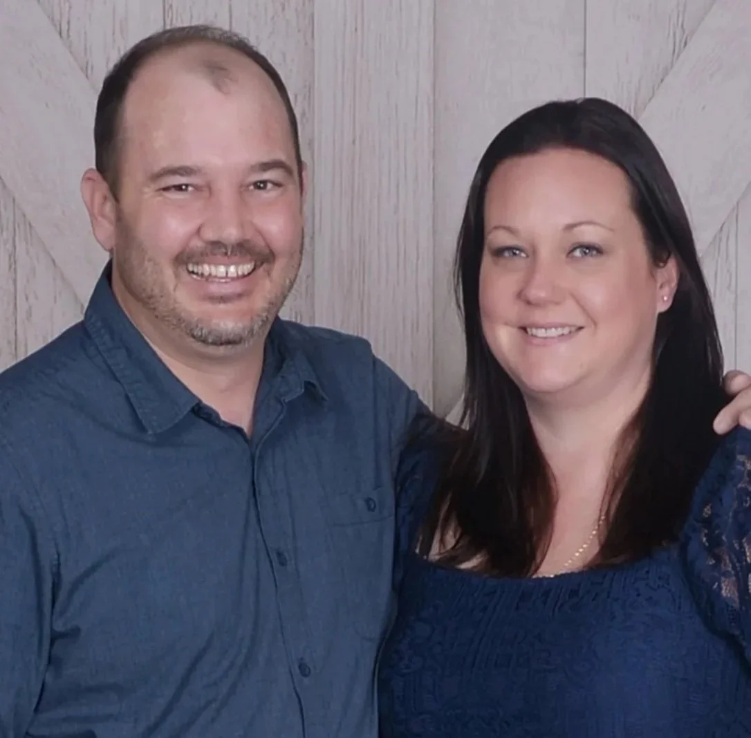 A man and a woman smiling, standing close together against a wooden wall background.