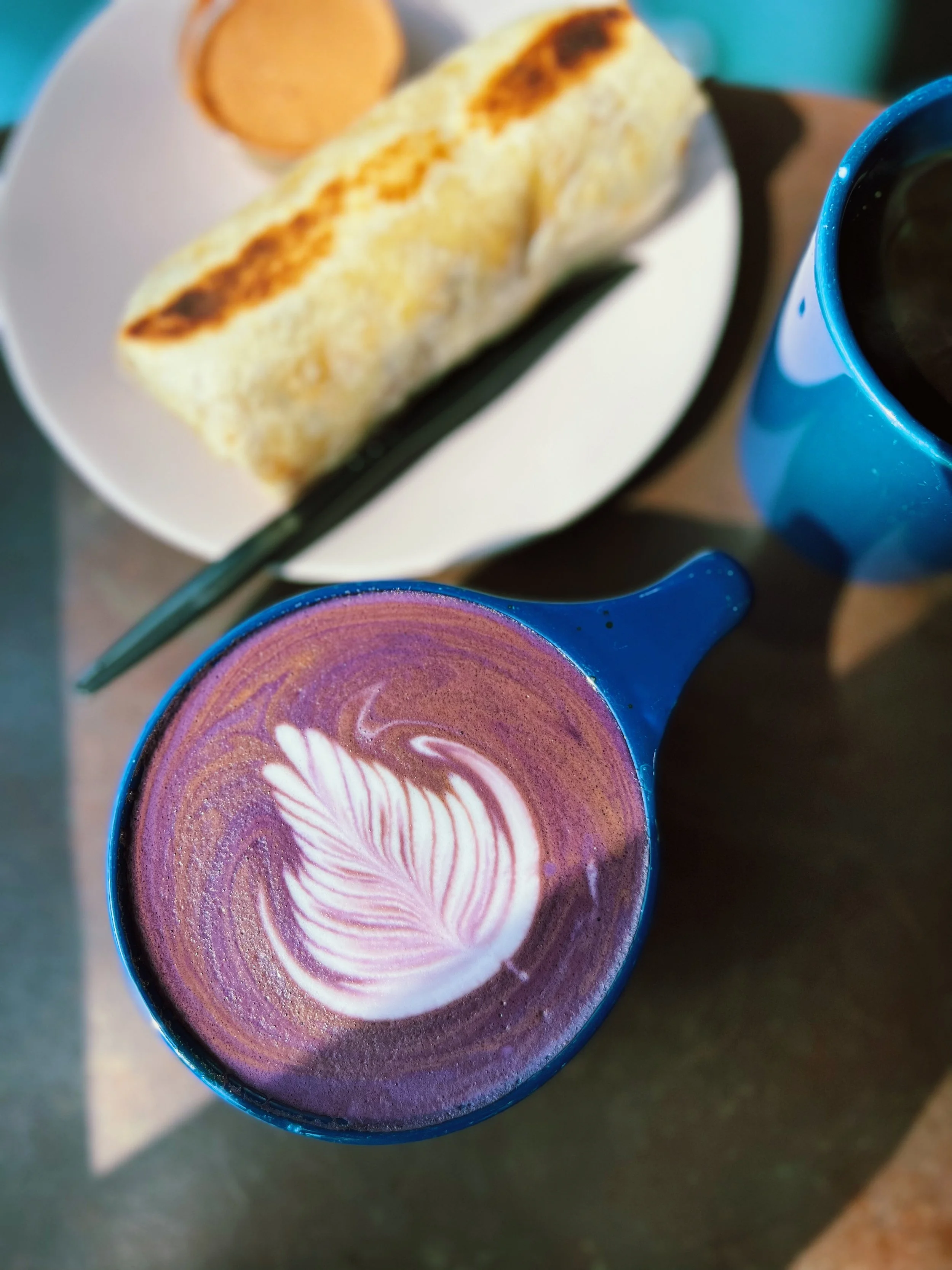 A blue mug filled with a purple and pink latte art drink, accompanied by a white plate with a toasted sandwich and a small bowl of sauce, and a black utensil.