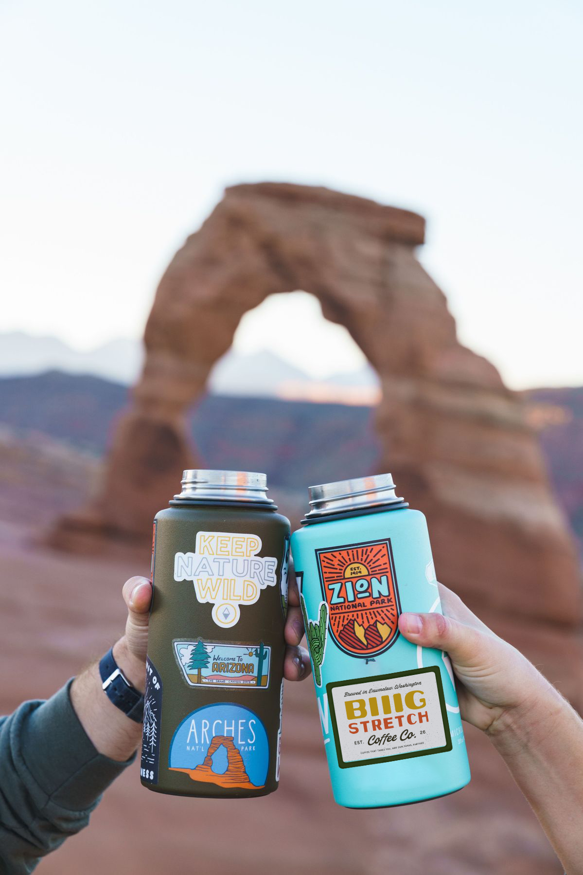 Two hands holding travel water bottles with stickers on them in front of Delicate Arch at Arches National Park with mountains in the background.