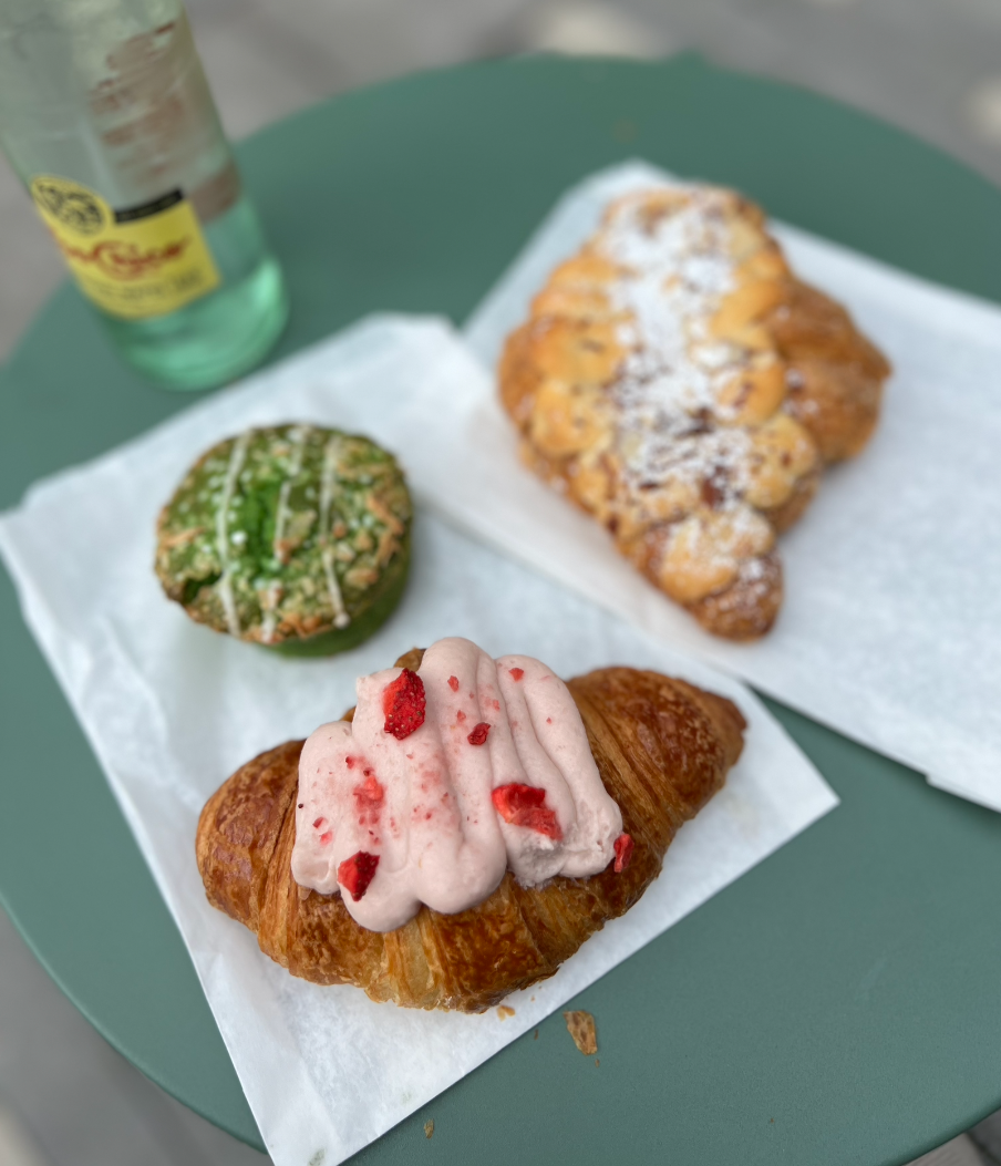 A green plate with a white napkin holding a croissant topped with pink whipped cream and red fruit pieces, a green glazed muffin with white drizzle, and a fried pastry dusted with powdered sugar. A bottle of sparkling water is partially visible in the background.